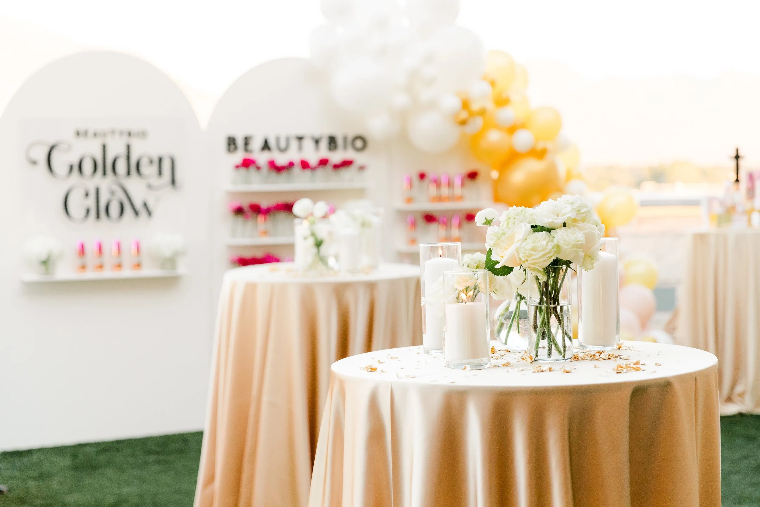 Decorative table with white flowers, candles, and petals, set up for an event in a bright, modern space with balloons and signage.