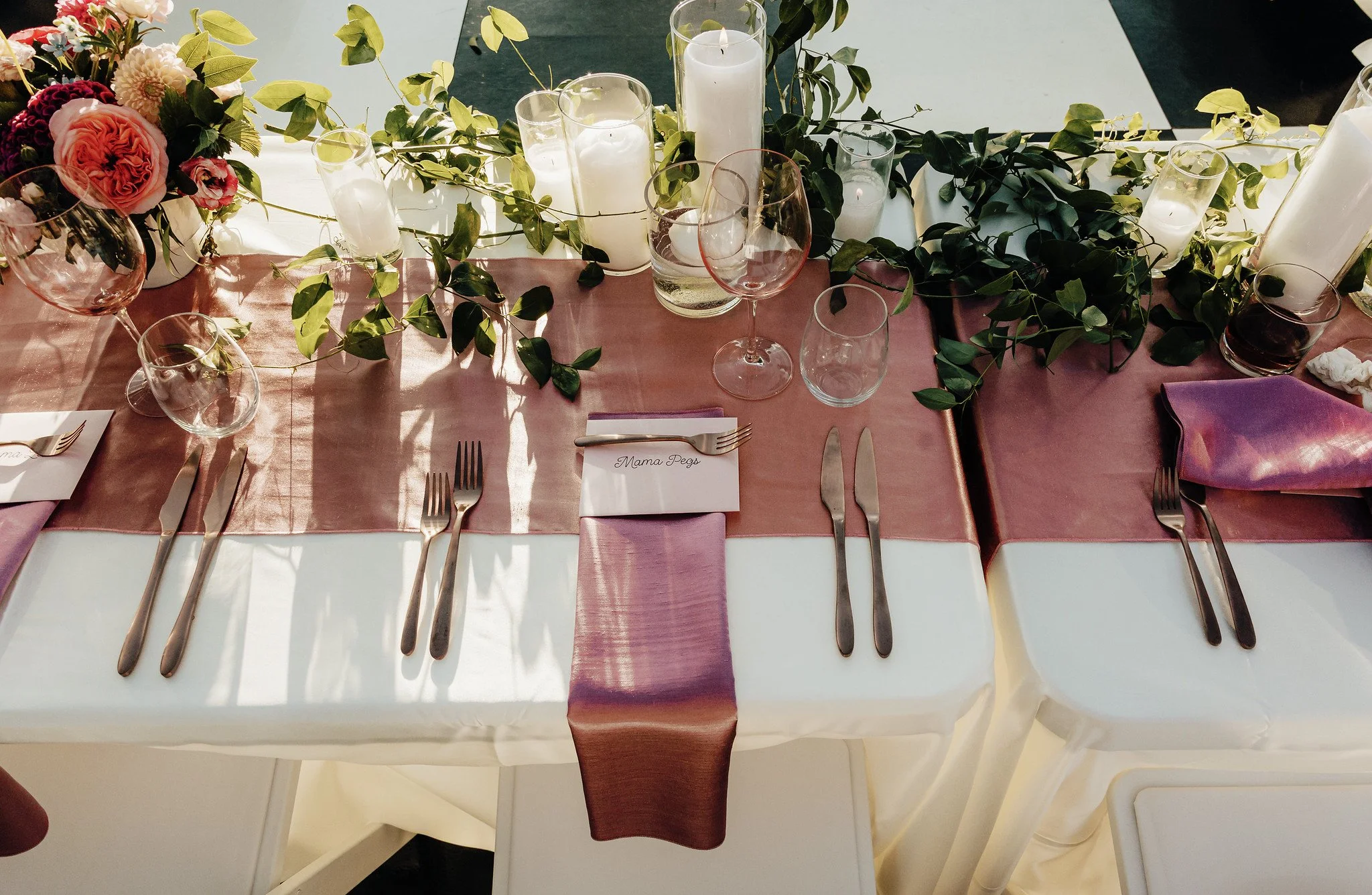 A decorated table with pink and white flowers, candles in glass holders, greenery, wine glasses, and neatly folded napkins for a festive occasion.