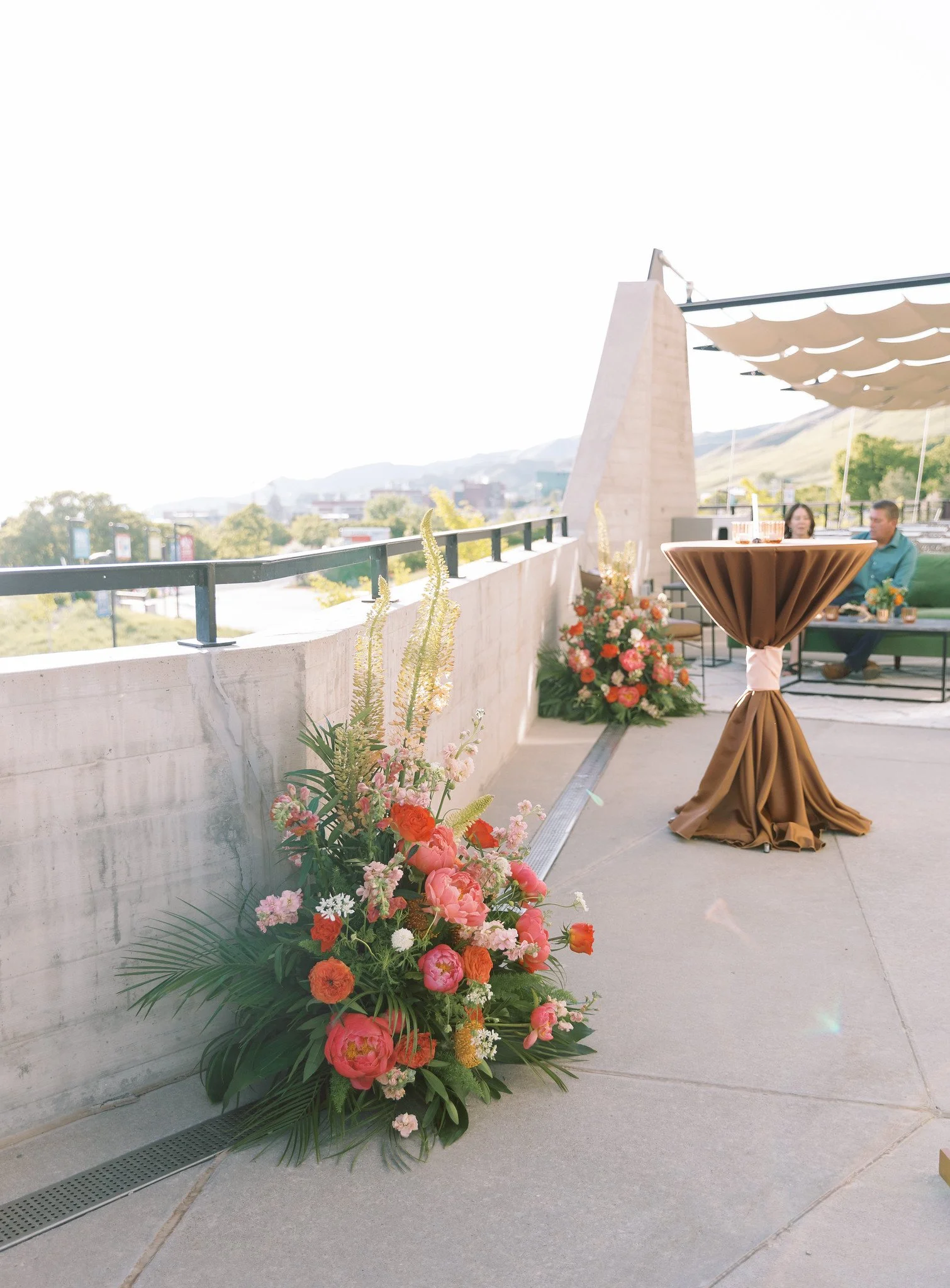 Outdoor terrace with floral arrangement, a round table with a brown tablecloth, and two people sitting and talking, overlooking a cityscape with mountains in the background.