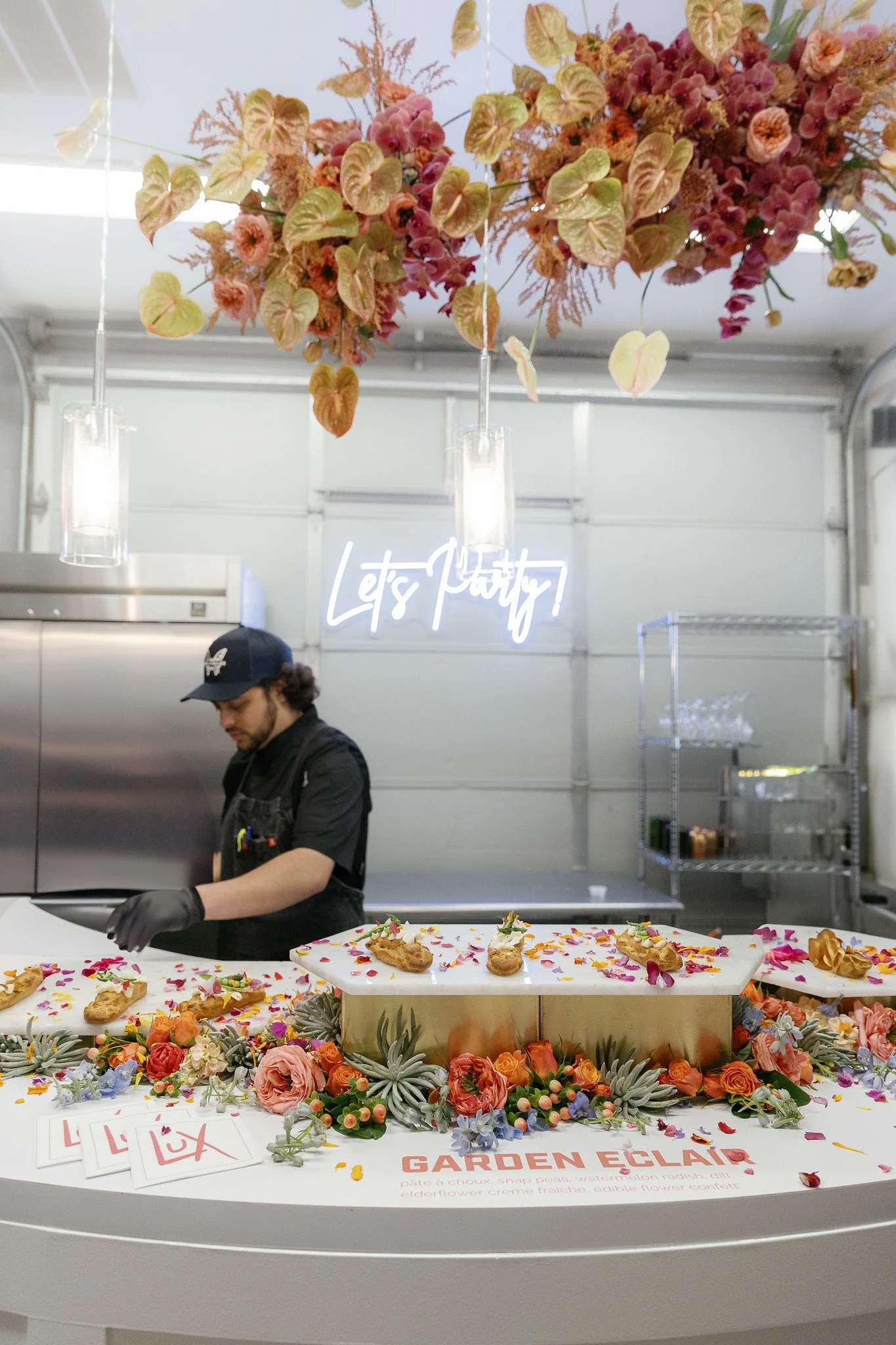A person wearing a black uniform and cap working at a dessert table decorated with colorful flowers. The table has a sign that says 'Garden Eclairs' and features éclairs decorated with flowers and petals.