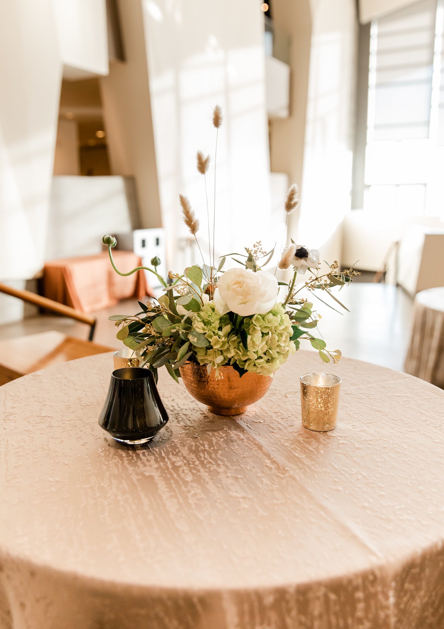 A floral arrangement with white roses, greenery, and beige pampas grass in a copper vase, placed on a textured beige table, with black and gold decorative cups beside it.