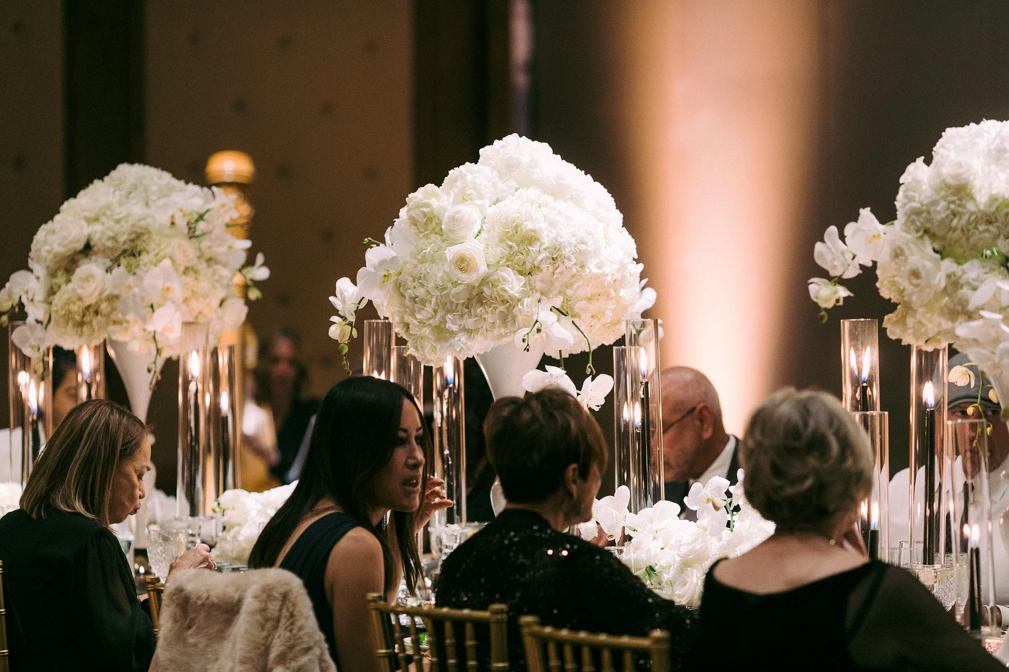 Elegant event with tall floral centerpieces featuring white flowers, surrounded by guests seated at tables with candles, in a dimly lit venue.
