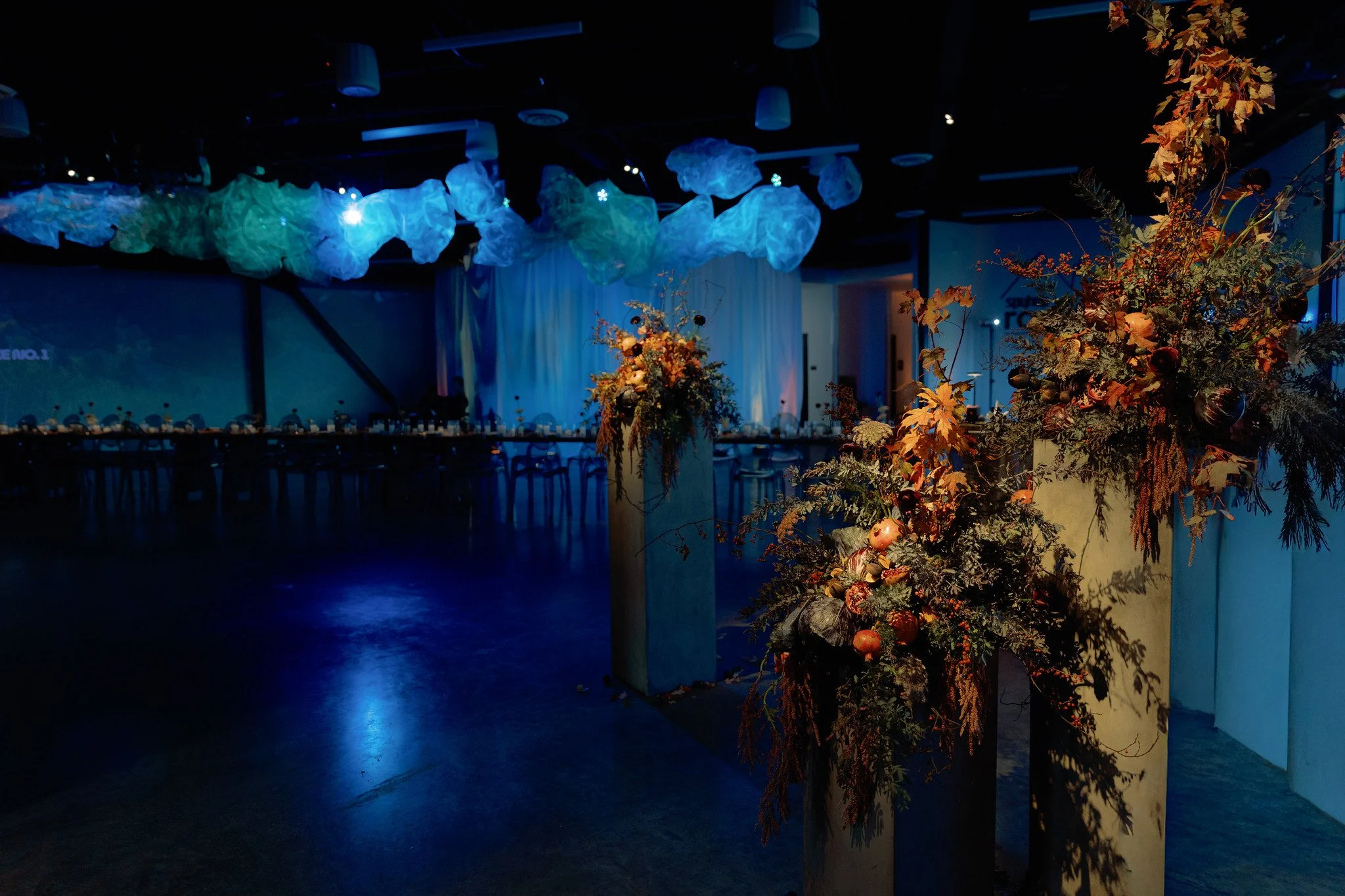 Indoor event space decorated with hanging cloud-like light fixtures and large floral arrangements with autumn-colored leaves, pumpkins, and dark foliage.