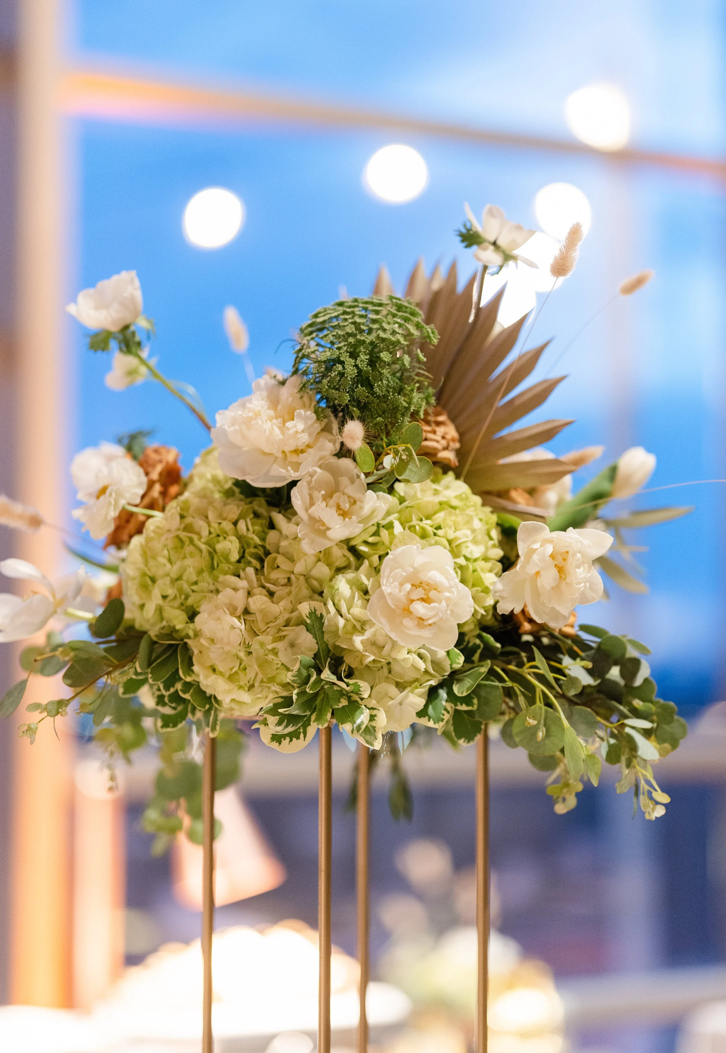 Close-up of a floral arrangement with white and green flowers, dried leaves, and foliage, with a soft background of blurred lights and sky.