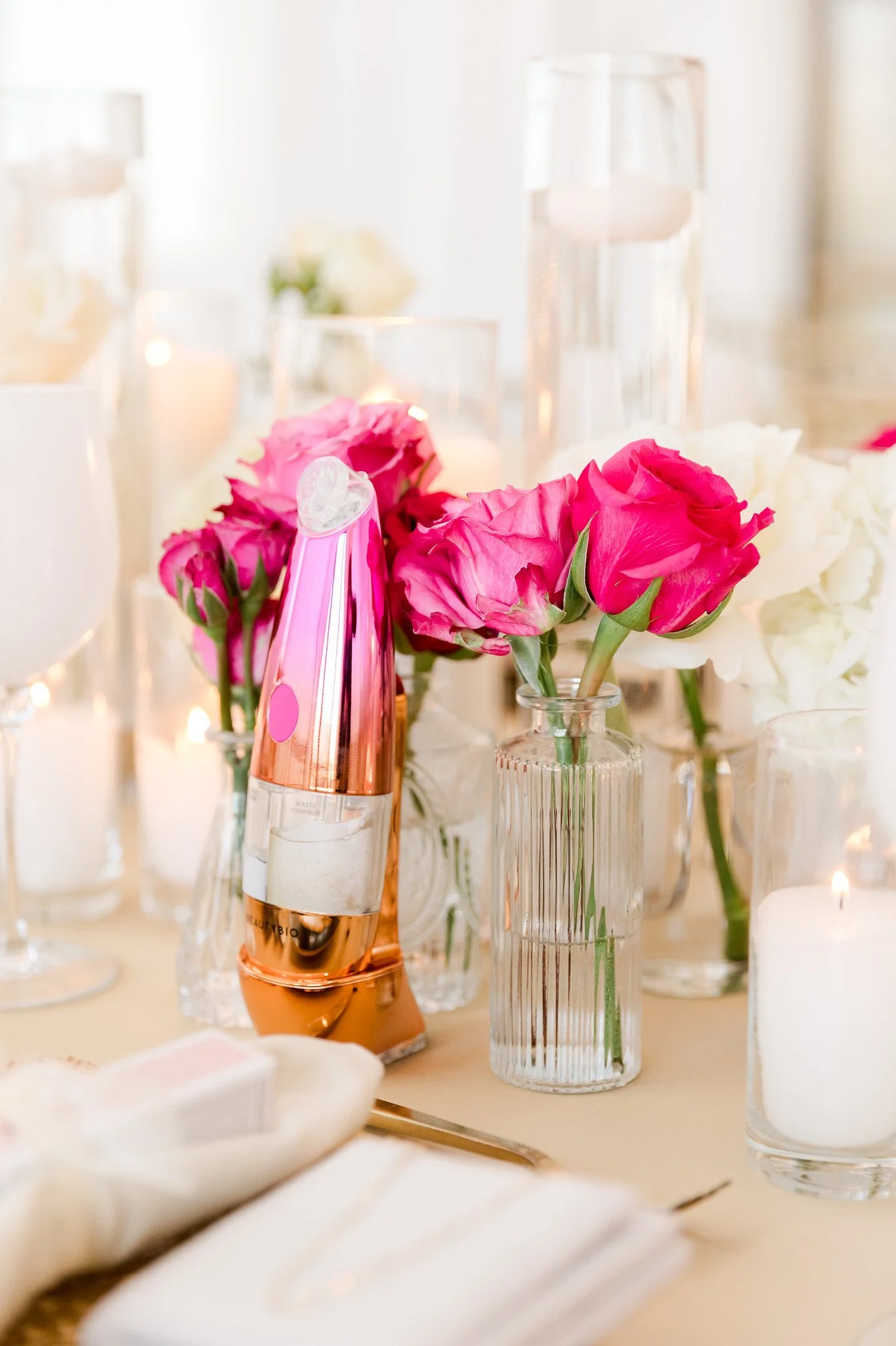 Close-up of pink and white roses in glass vases on a table, with candles and decorative items in the background, at a light-colored party or wedding table setting.