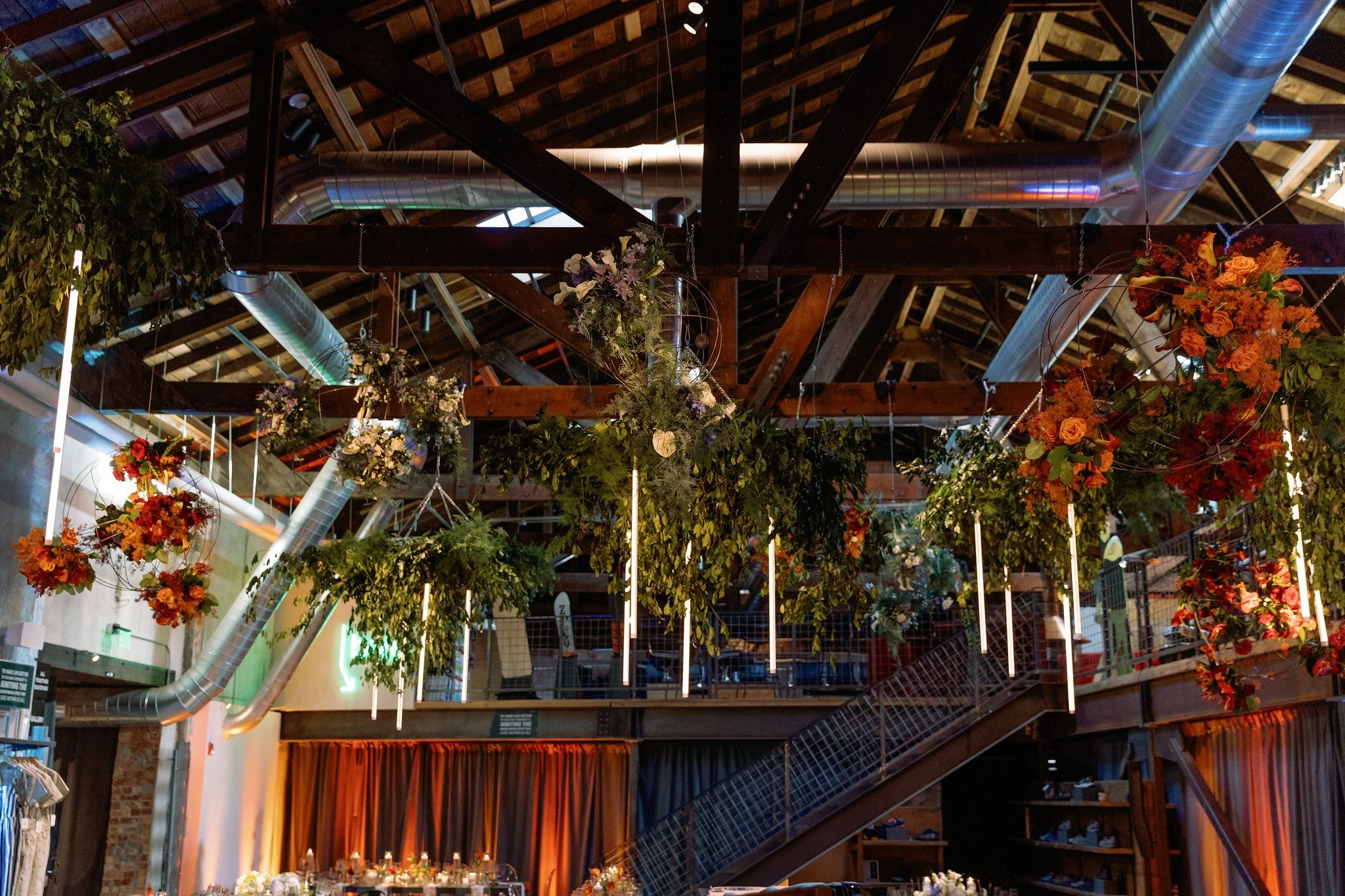 Indoor event space with floral arrangements hanging from the ceiling, exposed wooden beams, and large metal ventilation ducts.