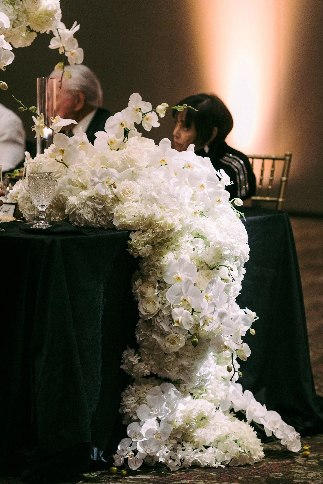 A wedding reception table decorated with a large white floral centerpiece of roses and orchids, with a woman and an older man sitting behind it, in a dimly lit room.