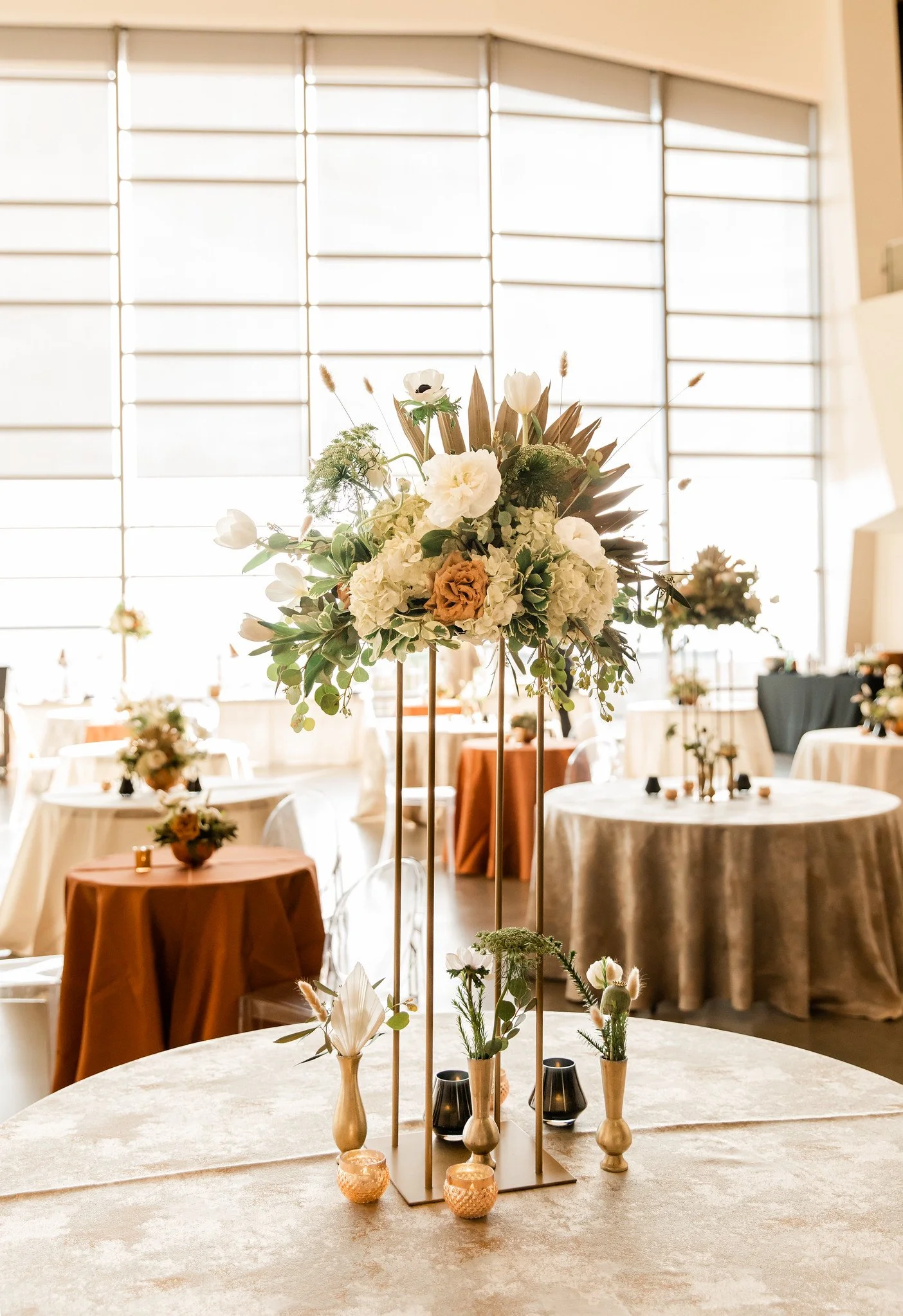 Elegant floral centerpiece with white and beige flowers and greenery on a high table, with decorated tables and large windows in a bright event space.