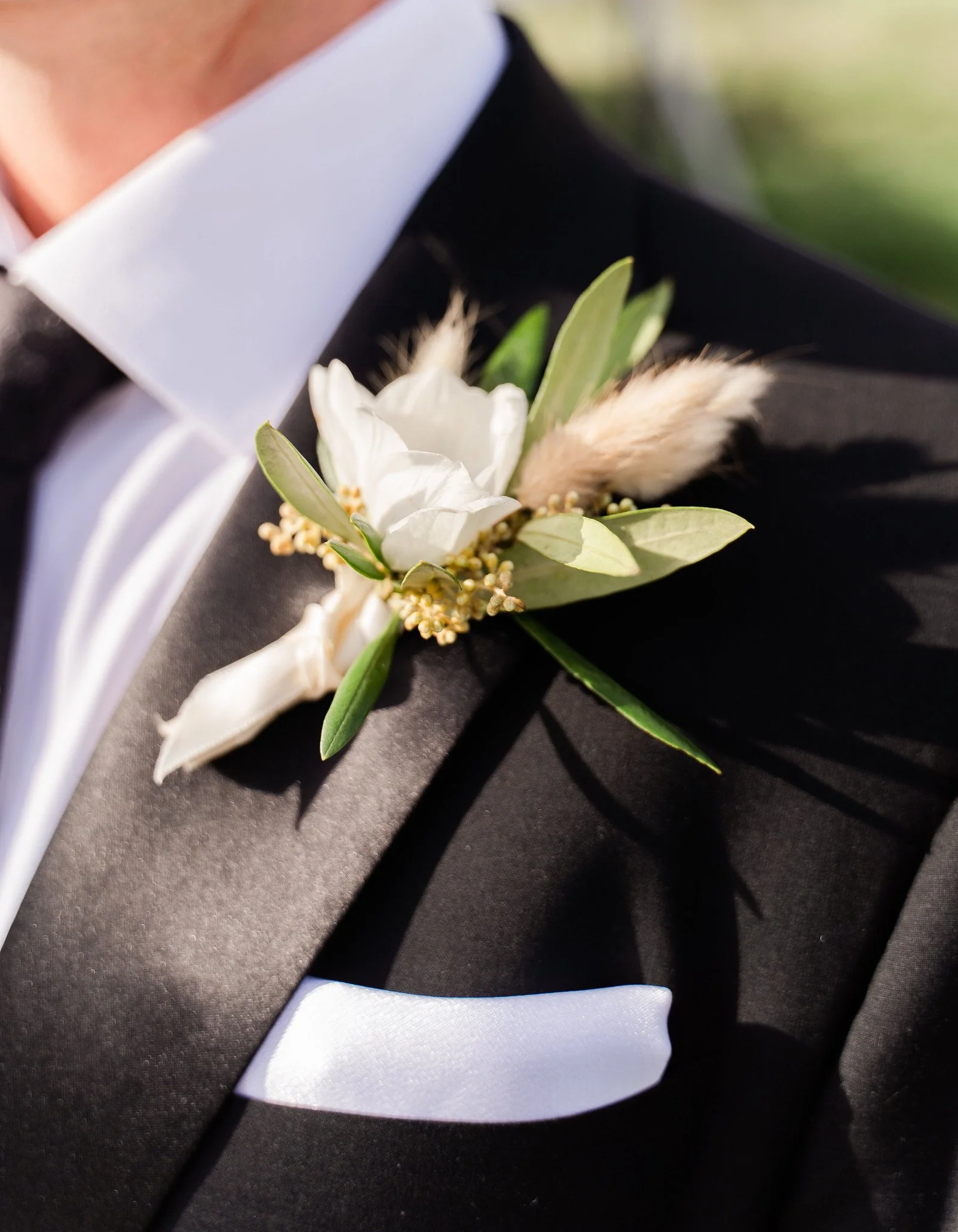 Close-up of a person wearing a black tuxedo with a white shirt and a boutonnière on the lapel, consisting of white flowers, green leaves, and a feathery accent, likely at a formal event or wedding.