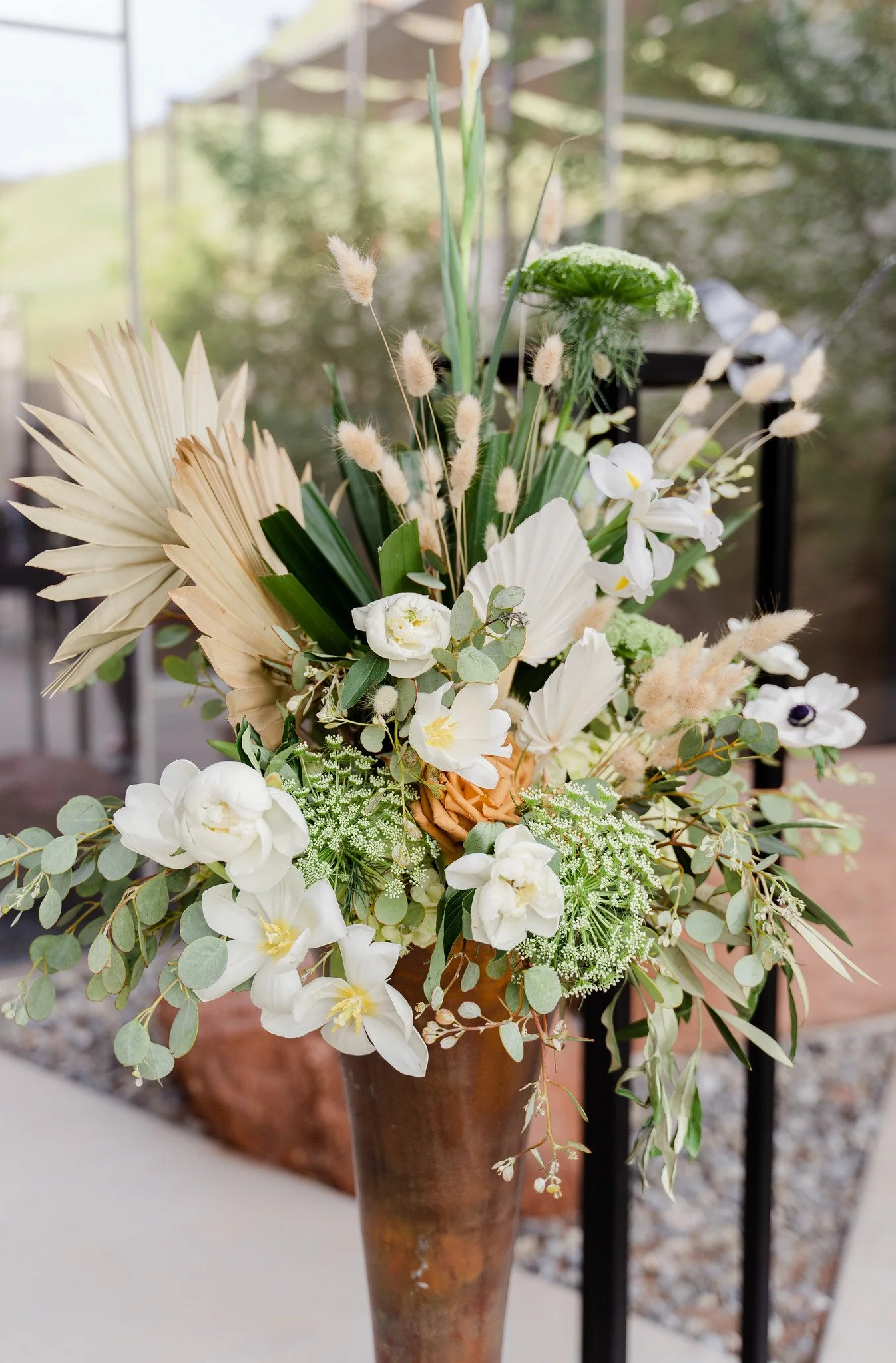 A tall flower arrangement in a copper vase with white flowers, green foliage, and dried elements, placed outdoors with a blurred background.