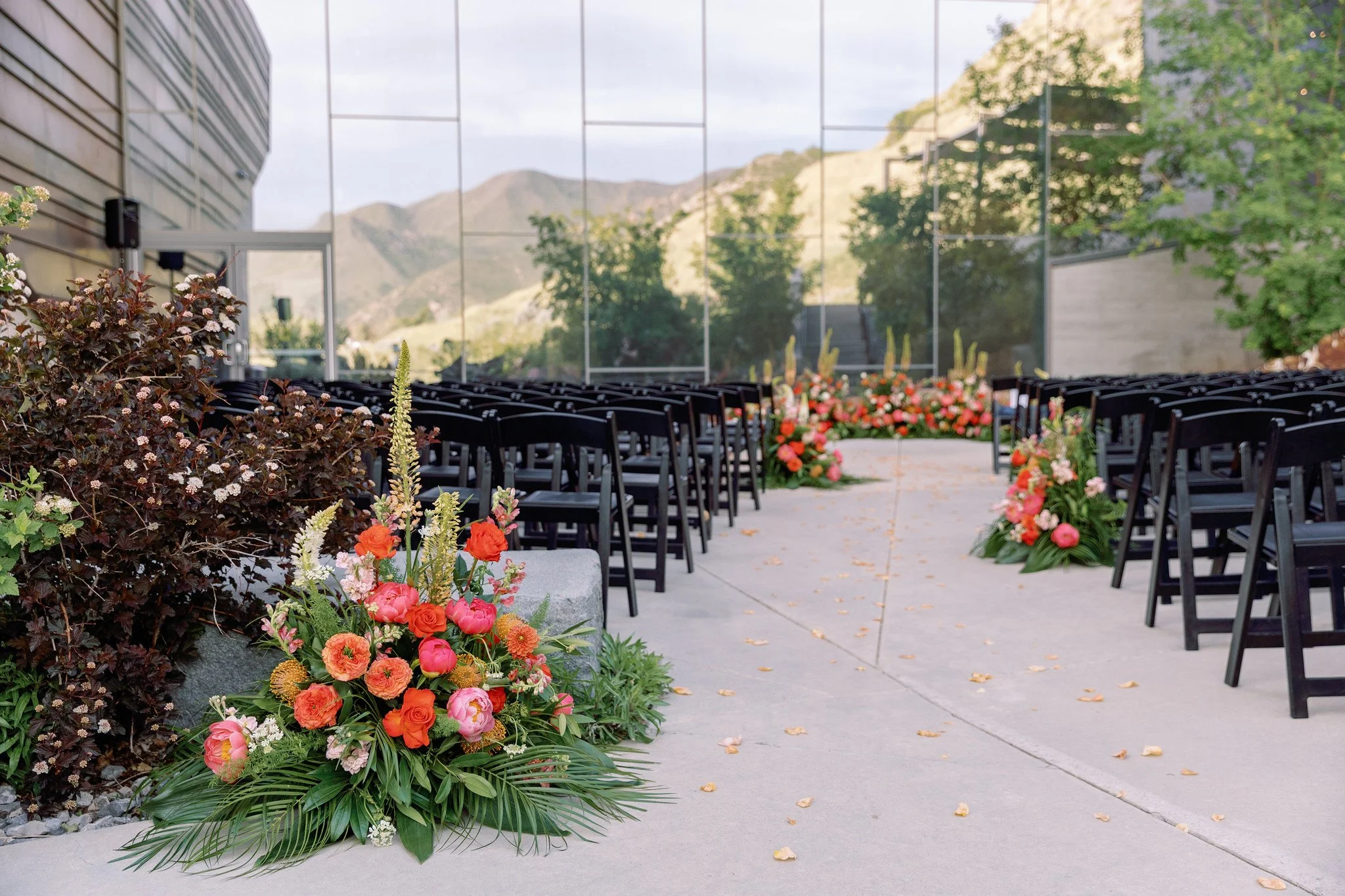 Outdoor wedding ceremony setup with black chairs, floral arrangements, and mountain view reflected in a glass wall.