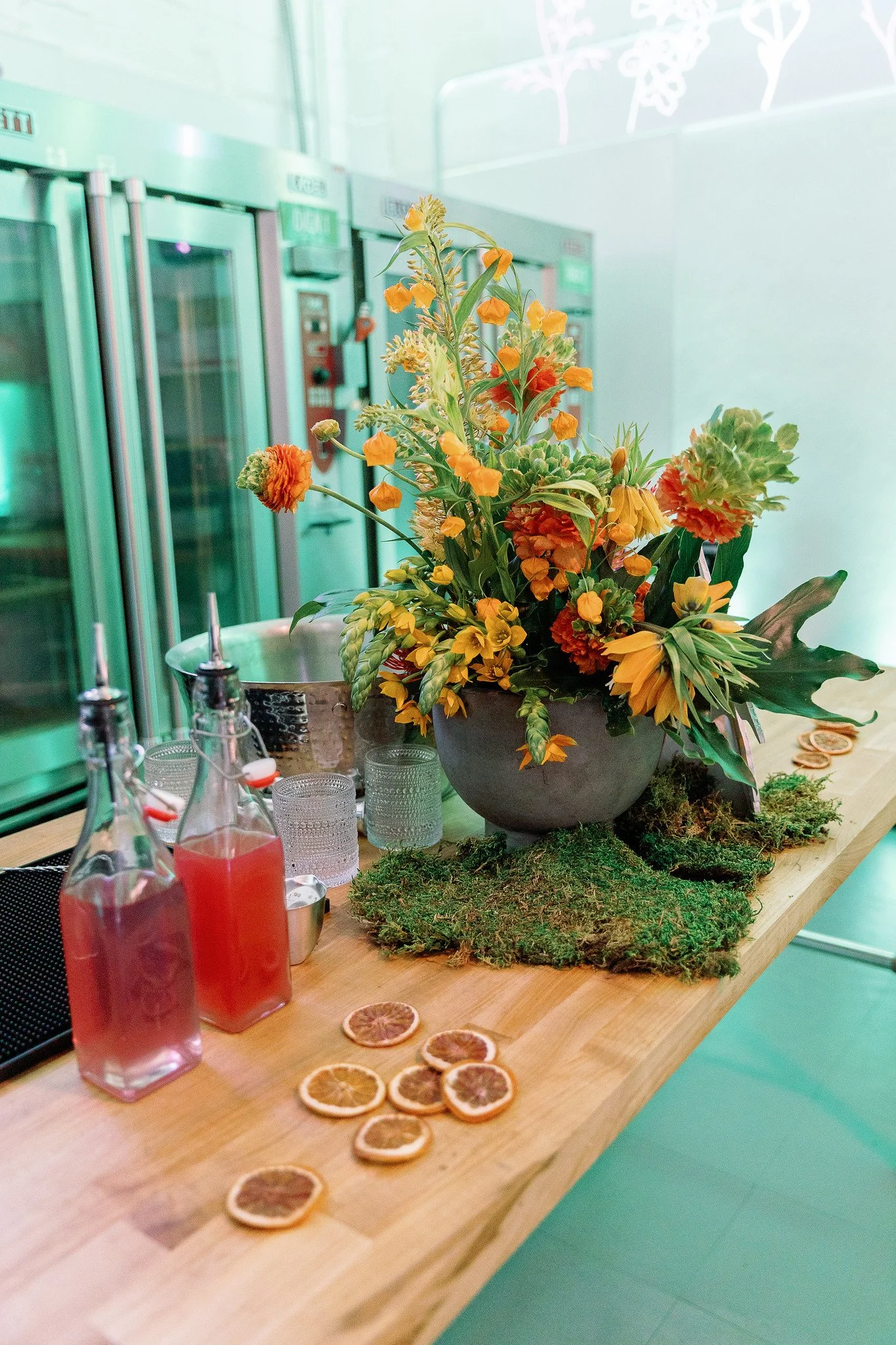 A floral arrangement with orange and yellow flowers in a gray pot on a wooden table, alongside bottles with red drinks, clear glasses, and dried orange slices, set against a background of green-colored kitchen appliances and wall.