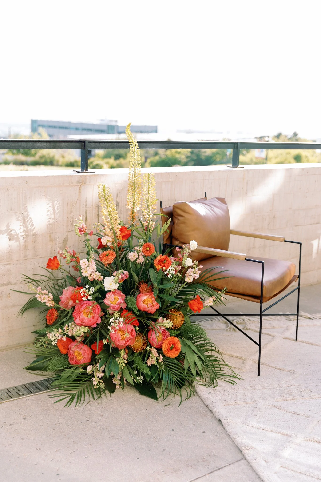 A large colorful flower arrangement featuring pink, orange, and cream flowers with green foliage, placed beside a brown leather chair on a concrete patio.