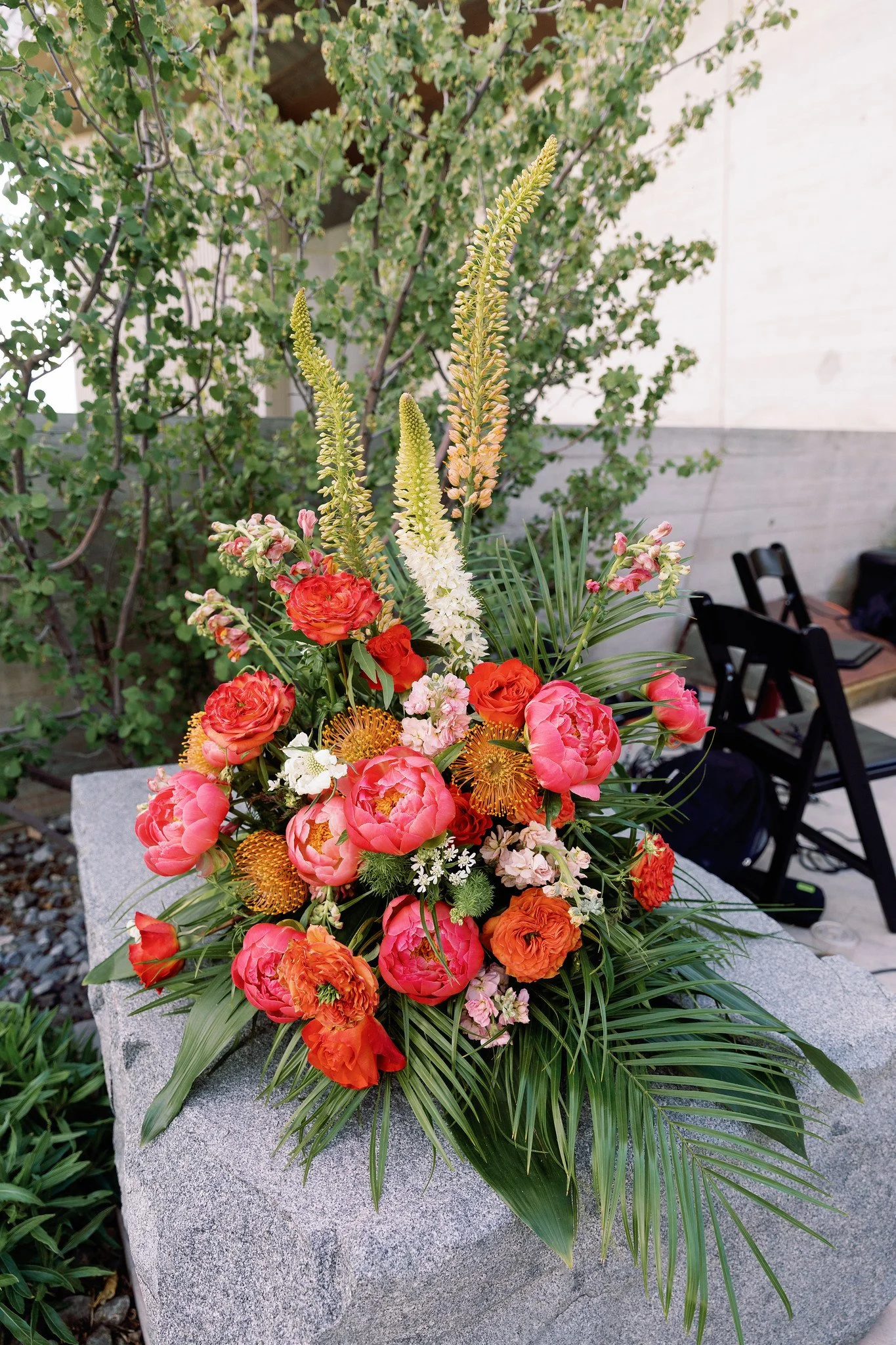 A colorful floral arrangement on a gray stone surface outdoors, including pink, orange, yellow, and white flowers with green foliage, in front of a blurred background of trees and chairs.