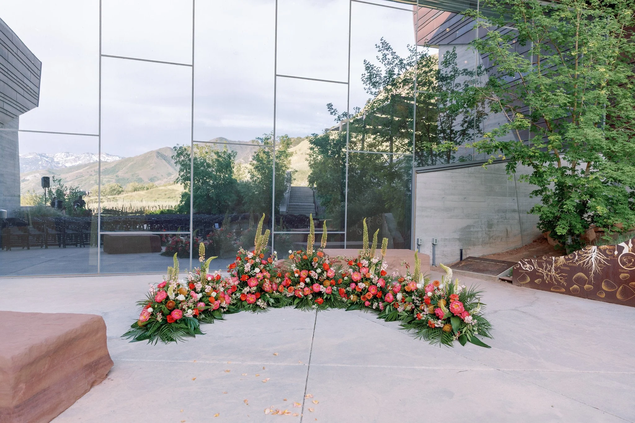 A large floral arrangement with pink and cream flowers and green foliage is placed outdoors on a paved area in front of a modern glass building. The building reflects trees and a mountain range with snow-capped peaks in the background.