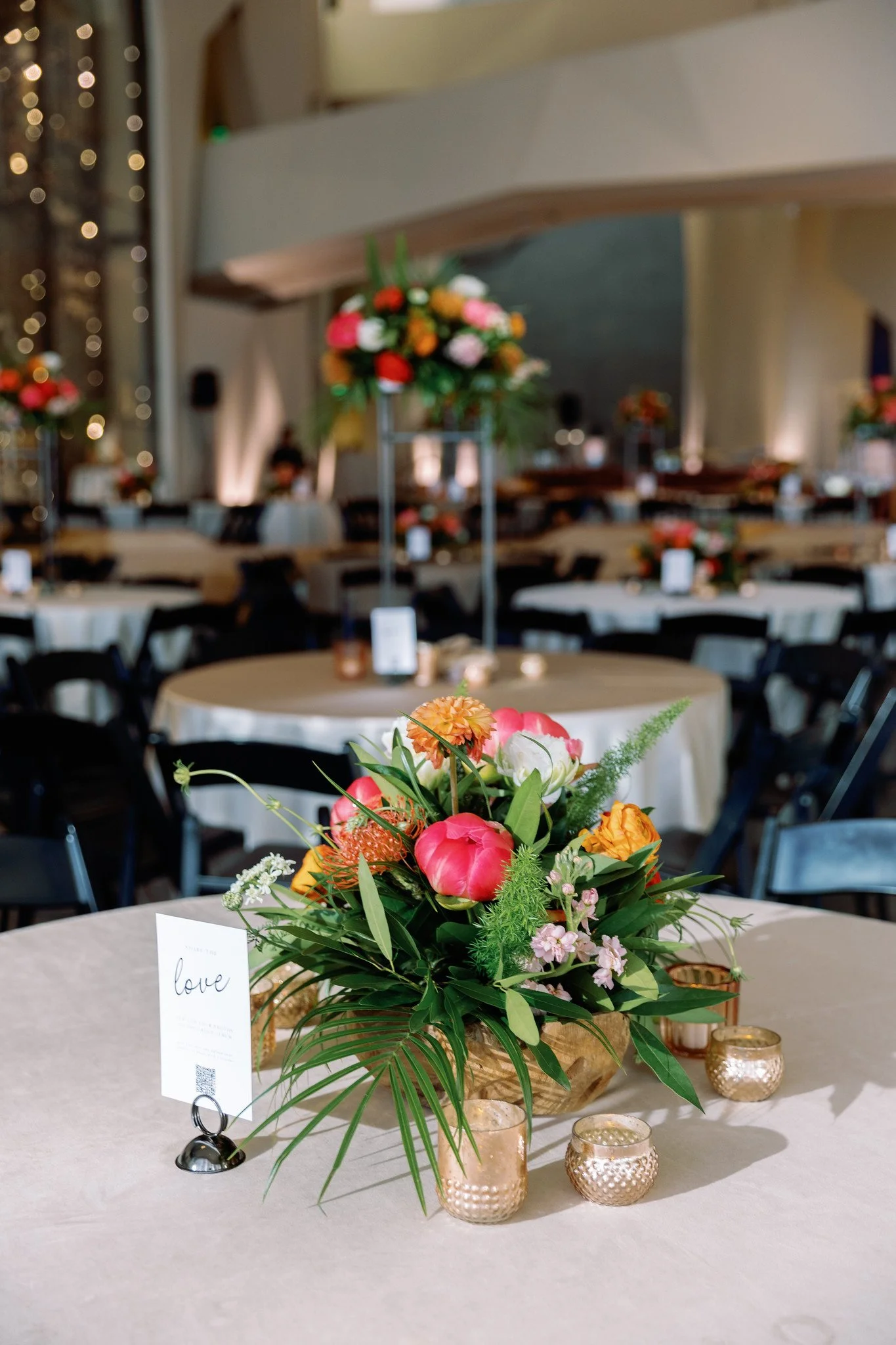 Floral arrangement with pink, orange, white, and green flowers on a table at a decorated event, with candles, place cards, and a blurred background of other tables and floral centerpieces.