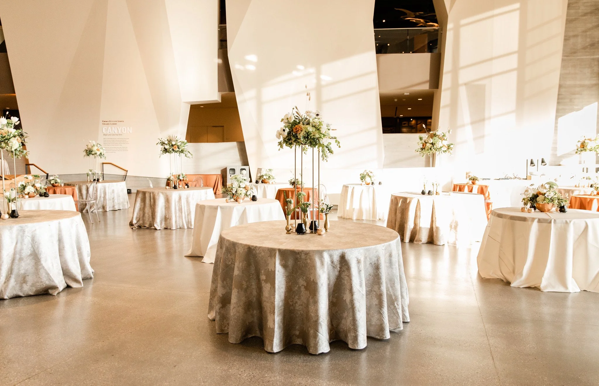 Event space with round tables covered in beige tablecloths and decorated with floral arrangements, modern architectural interior with large concrete wall, and sunlight casting geometric shadows.