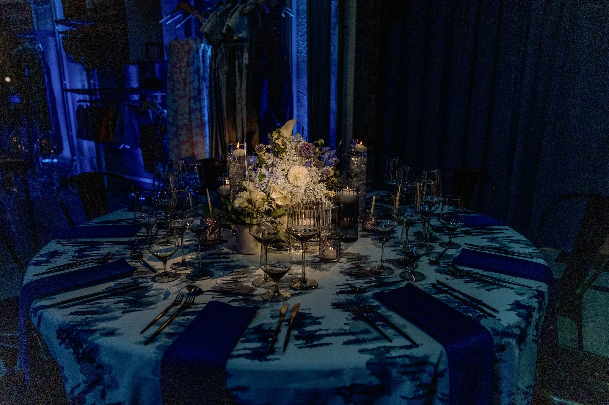 A round dining table set for an event, with a floral centerpiece, candles, and glassware, in a dimly lit setting with dark curtains and clothing hanging in the background.