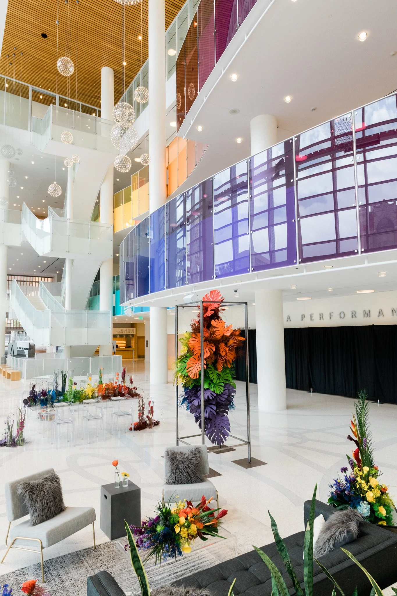 Interior of a modern building with colorful glass railings, hanging spherical light fixtures, and floral arrangements on display.