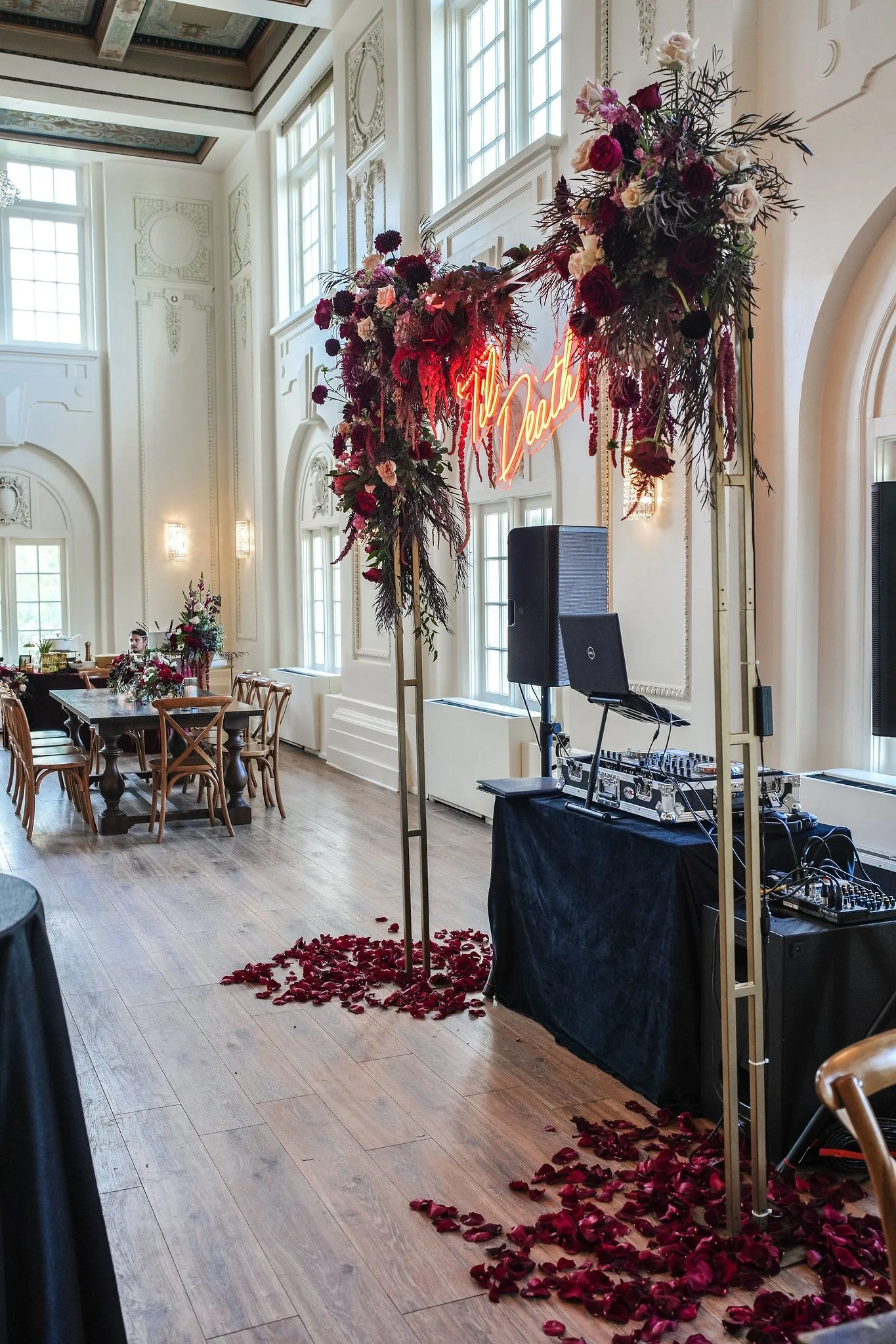 Elegant indoor venue decorated for a wedding, featuring a large flower arrangement with roses and greenery, a neon sign that reads 'You Love,' and a DJ setup with sound equipment. Rose petals are scattered on the wooden floor.