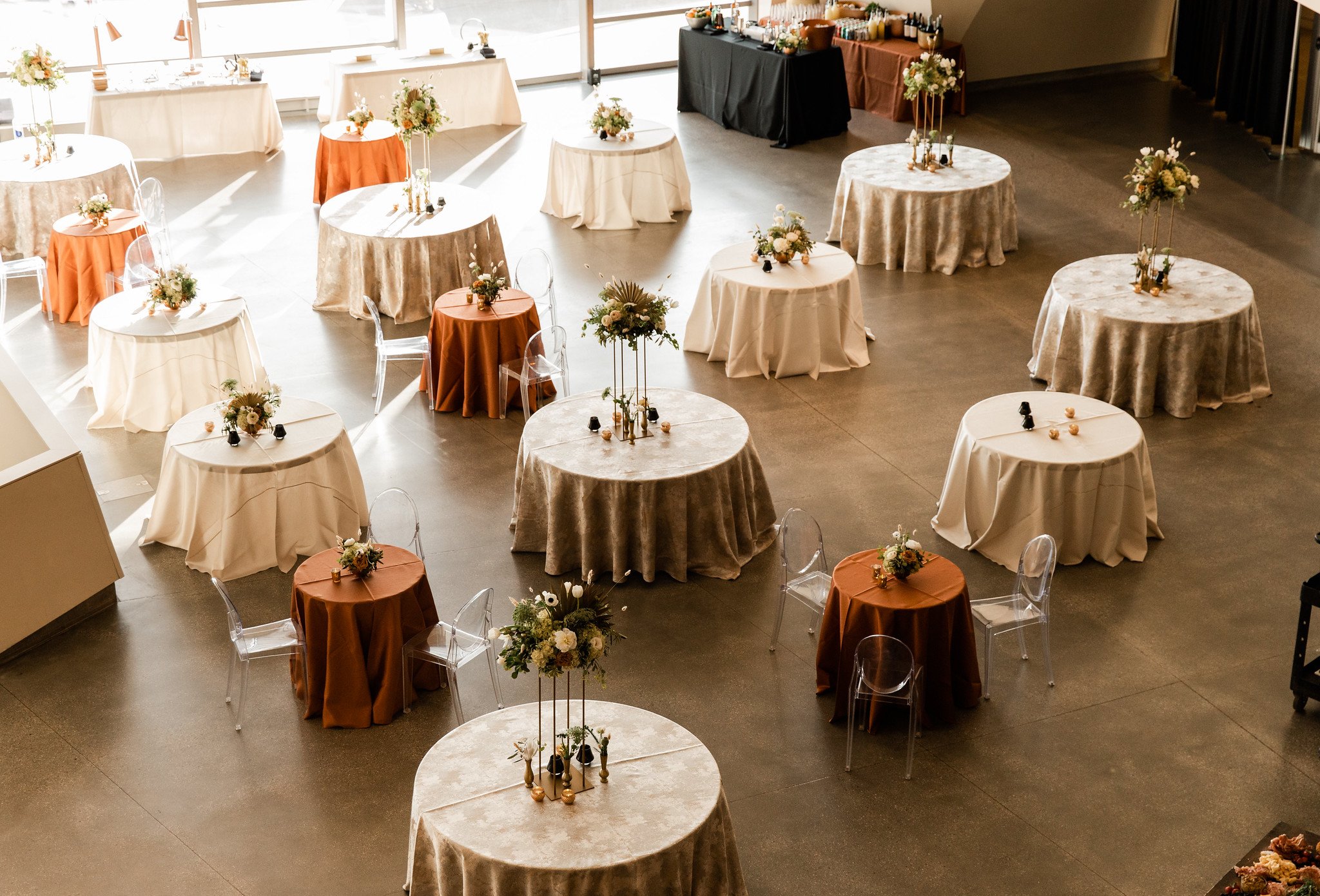 Elegant wedding or event reception setup with round tables covered in cream and brown tablecloths, each decorated with floral centerpieces, set in a spacious area with large windows and a dessert or drink station at the back.