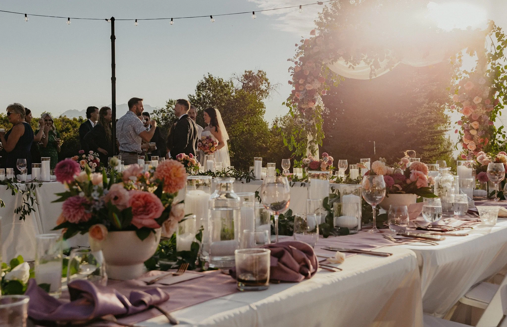 Wedding reception setup outdoors during sunset with decorated tables, floral centerpieces, and a floral arch with hanging lights in the background.