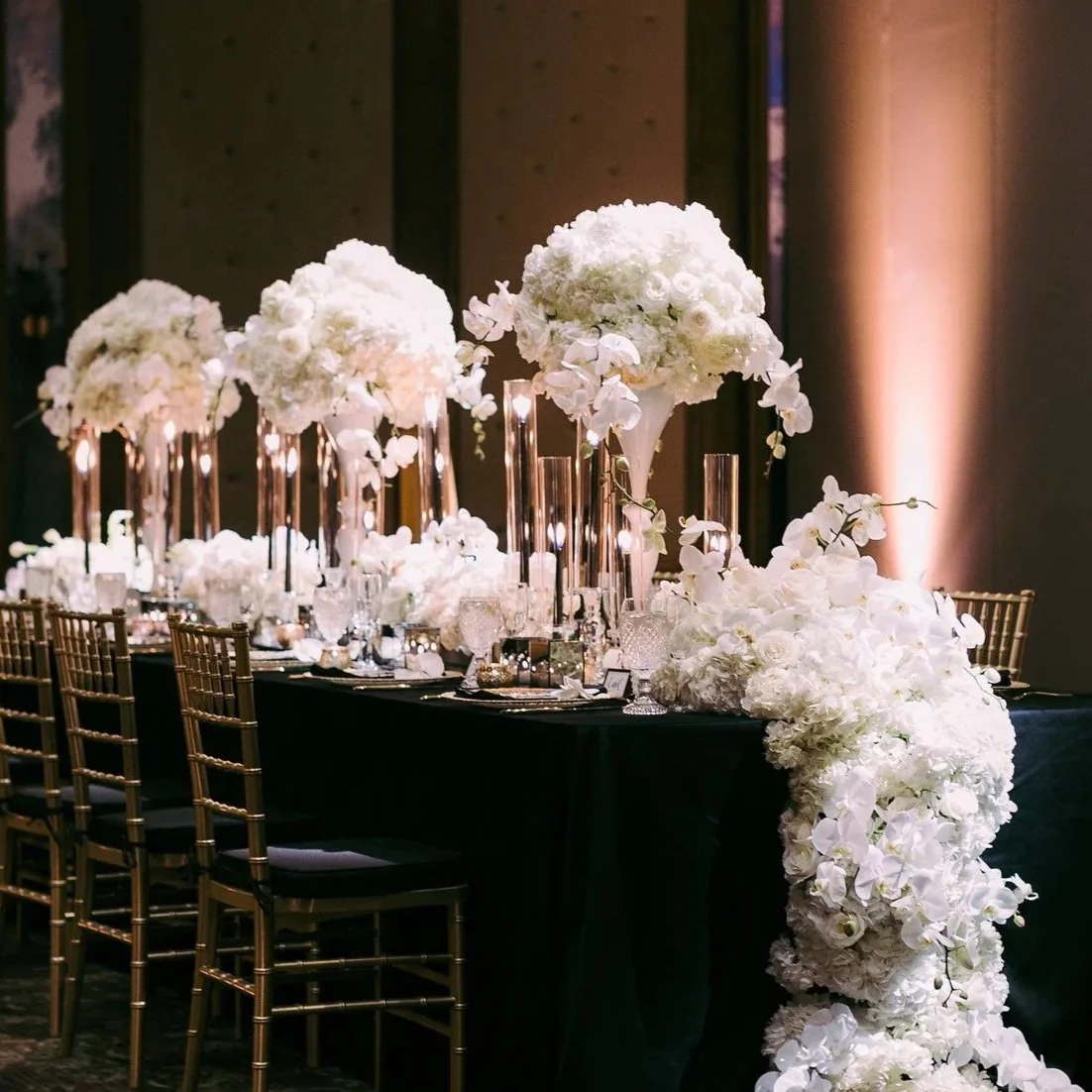 Elegant banquet table decorated with tall glass vases filled with white floral arrangements, including orchids and roses, set on a dark tablecloth with gold chairs.