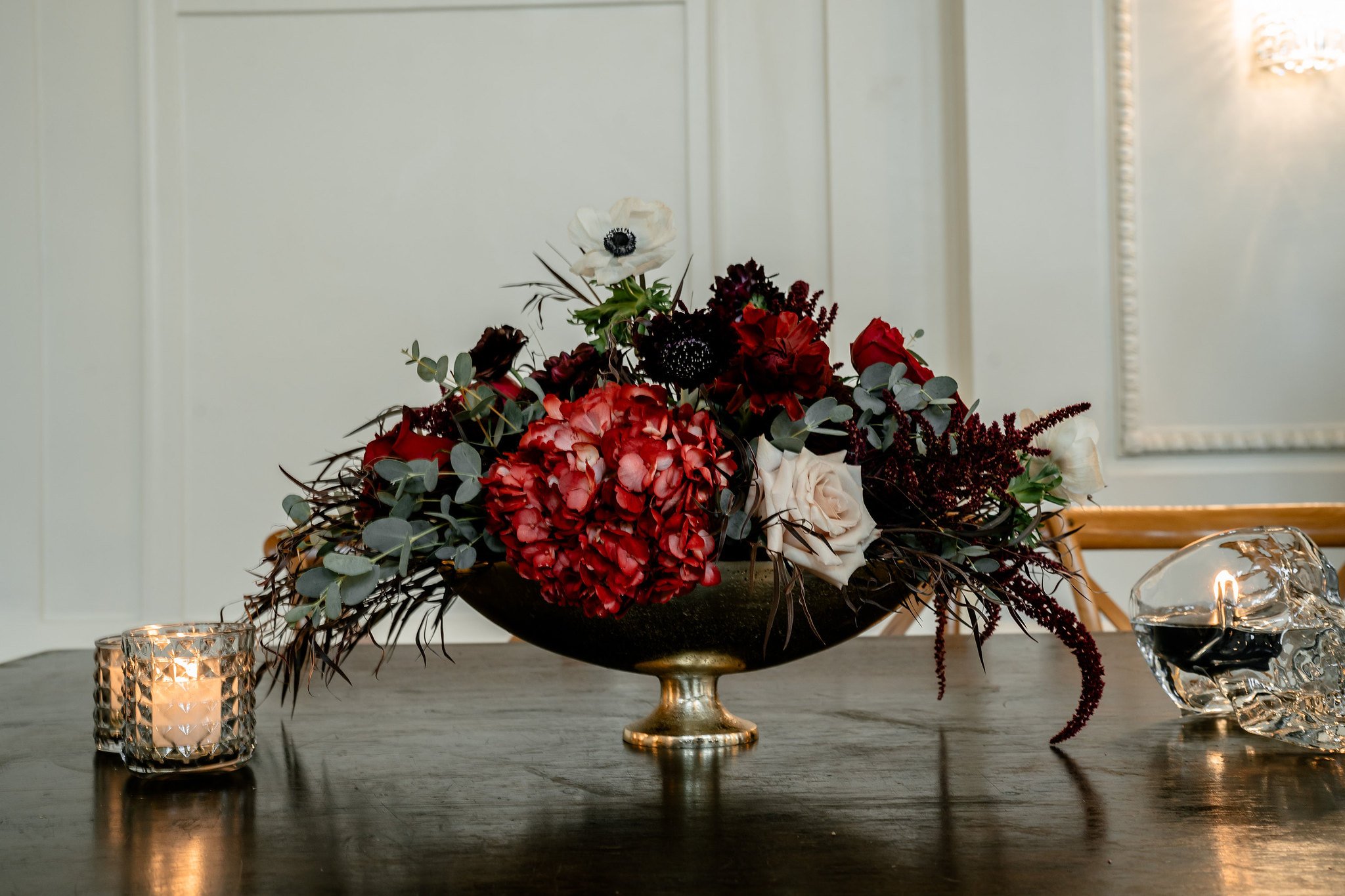 A floral centerpiece with red, white, and dark purple flowers, eucalyptus leaves, and decorative branches on a table, with a lit candle in a textured holder and a glass crystal skull nearby, against a white paneled wall.