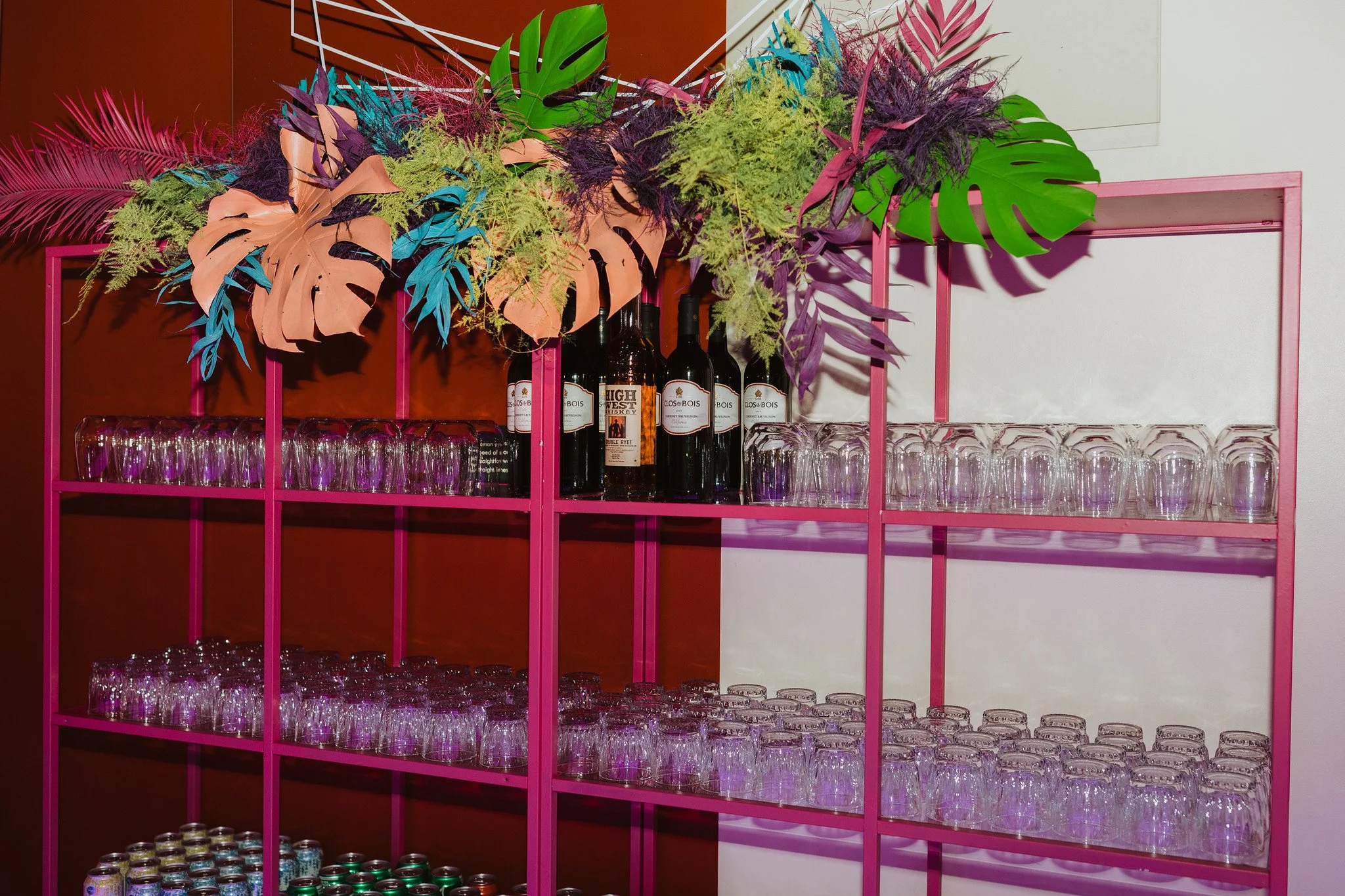 Pink metal shelf with empty glassware and bottles, decorated with a colorful arrangement of tropical leaves and plants including monstera leaves, ferns, and purple and pink foliage.