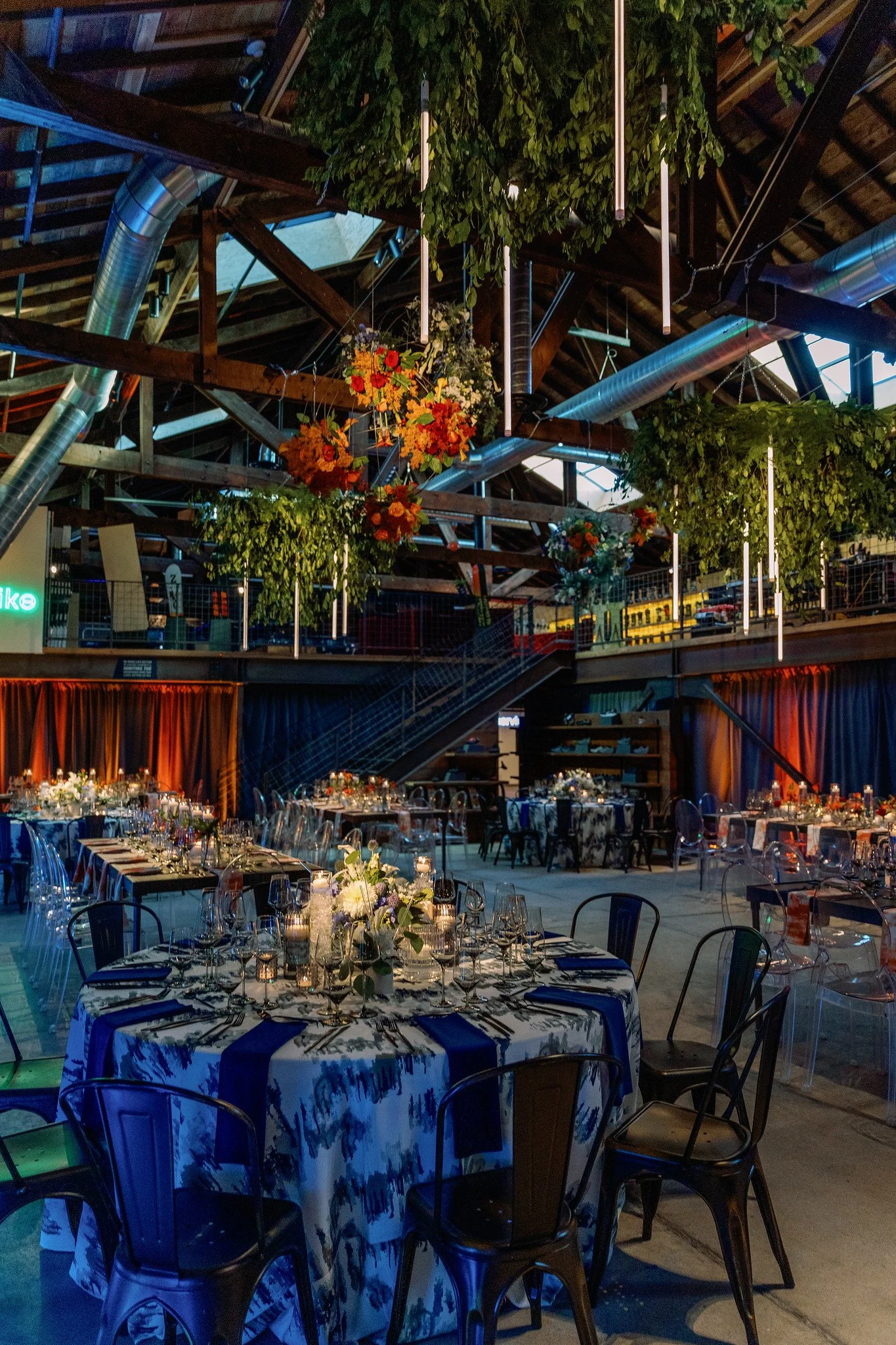 A large indoor space decorated for a formal event, with round tables covered in floral tablecloths and set with glassware and candles. Hanging from the ceiling are green plants and colorful floral arrangements, with visible metal ductwork and wooden 