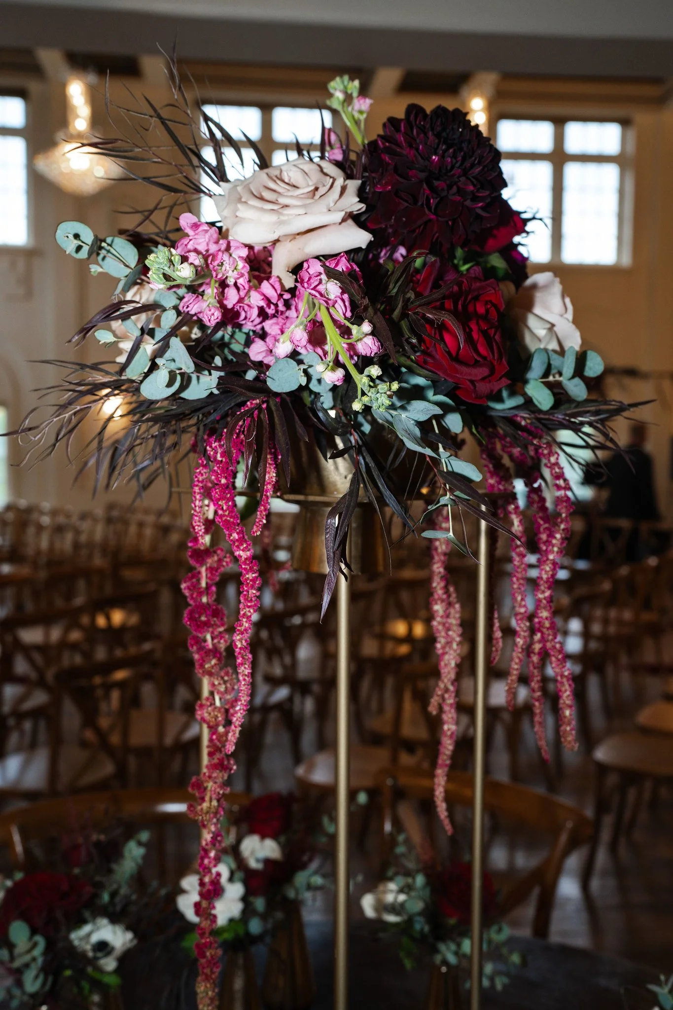A floral arrangement with white roses, dark red dahlias, pink flowers, greenery, and hanging pink amaranthus, set on a gold stand in a room with wooden chairs and large windows.
