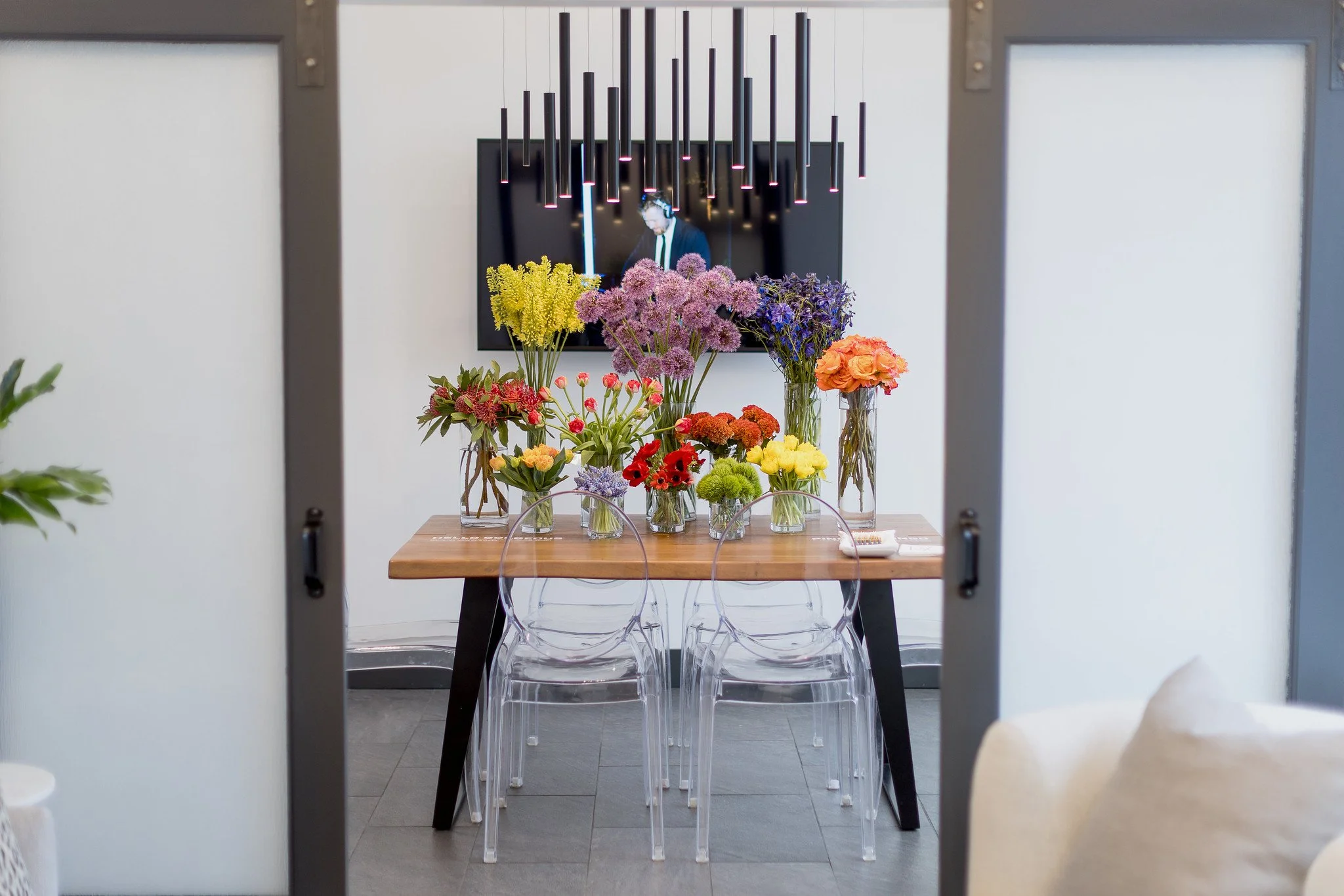 A dining table filled with various colorful flower arrangements in glass vases, viewed through a doorway with two white panels, in a modern space with gray flooring and white walls.