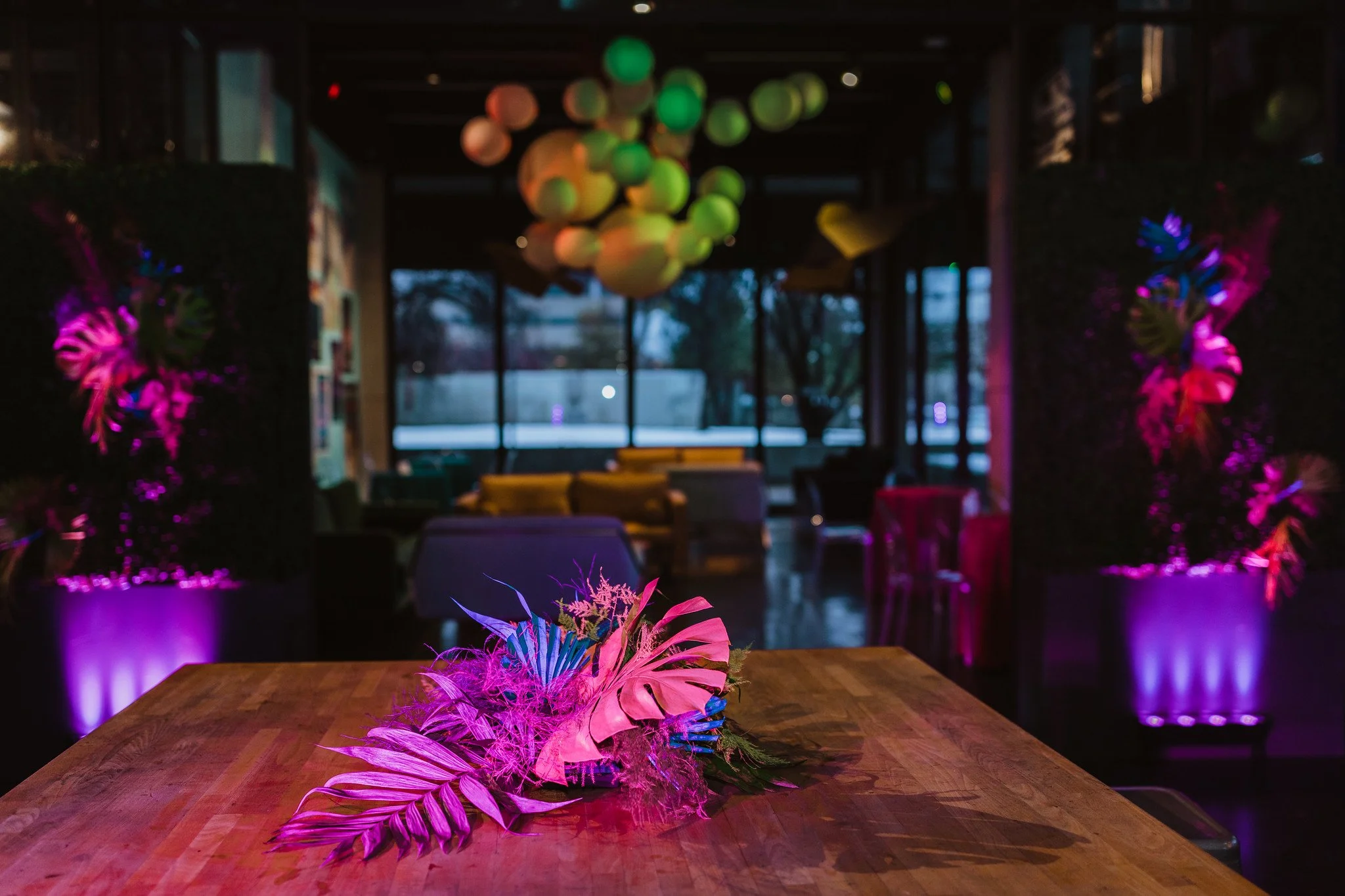 Decorative table centerpiece with vibrant pink and purple feathers, flowers, and foliage in a dimly lit event space with colorful lighting and modern decor.