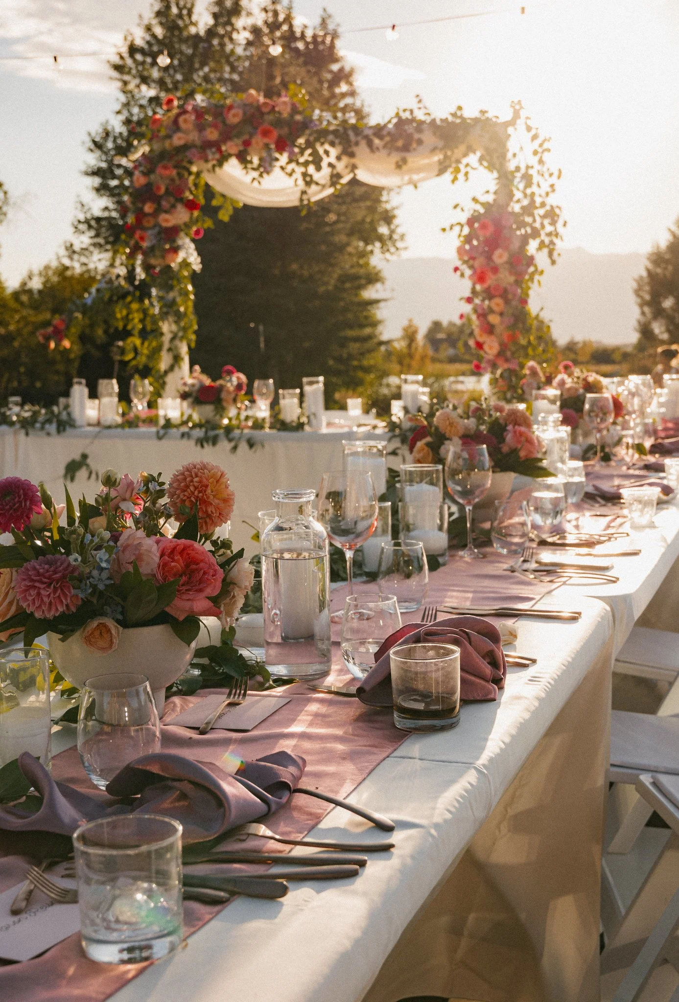A long outdoor dining table decorated with pink and purple flowers, lanterns, wine glasses, and cloth napkins, set up for a wedding or celebration in a garden during sunset.