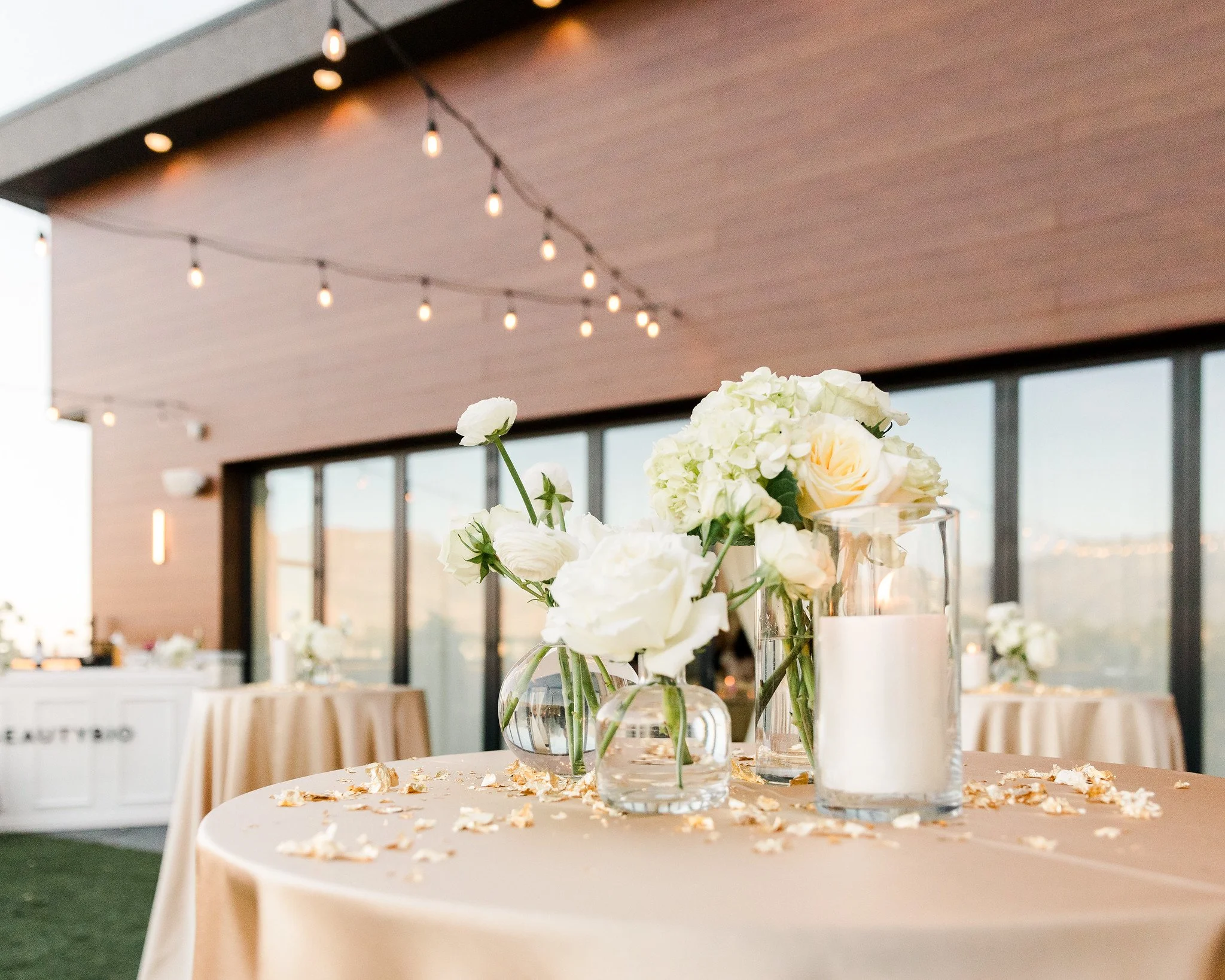 Decorative table with glass vases containing white and light peach flowers on a beige tablecloth at an indoor event space with string lights hanging from the ceiling.