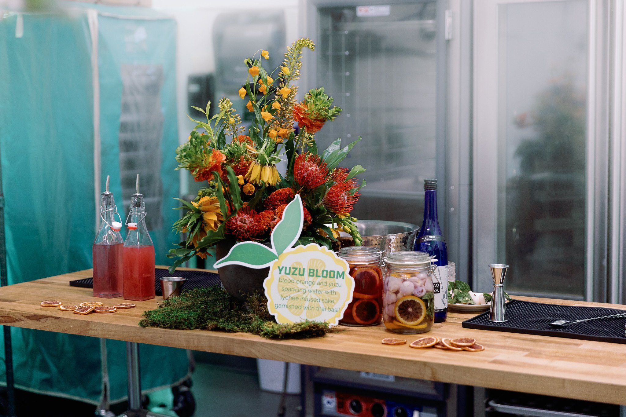 A colorful flower arrangement with a sign reading "Yuzu Bloom blood orange and yuzu sparkling water with lychee infused sake garnished with Thai basil" is placed on a wooden table. Nearby are bottles of pink liquid, jars of fruit slices, and dried citrus slices, suggesting a beverage station in a modern setting.