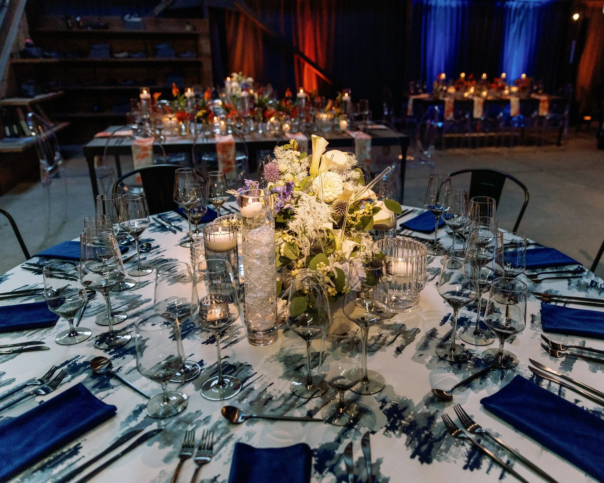 Elegant banquet table setup with floral centerpiece, candles, wine glasses, and navy blue napkins, in a dimly lit reception hall with decorated back wall.