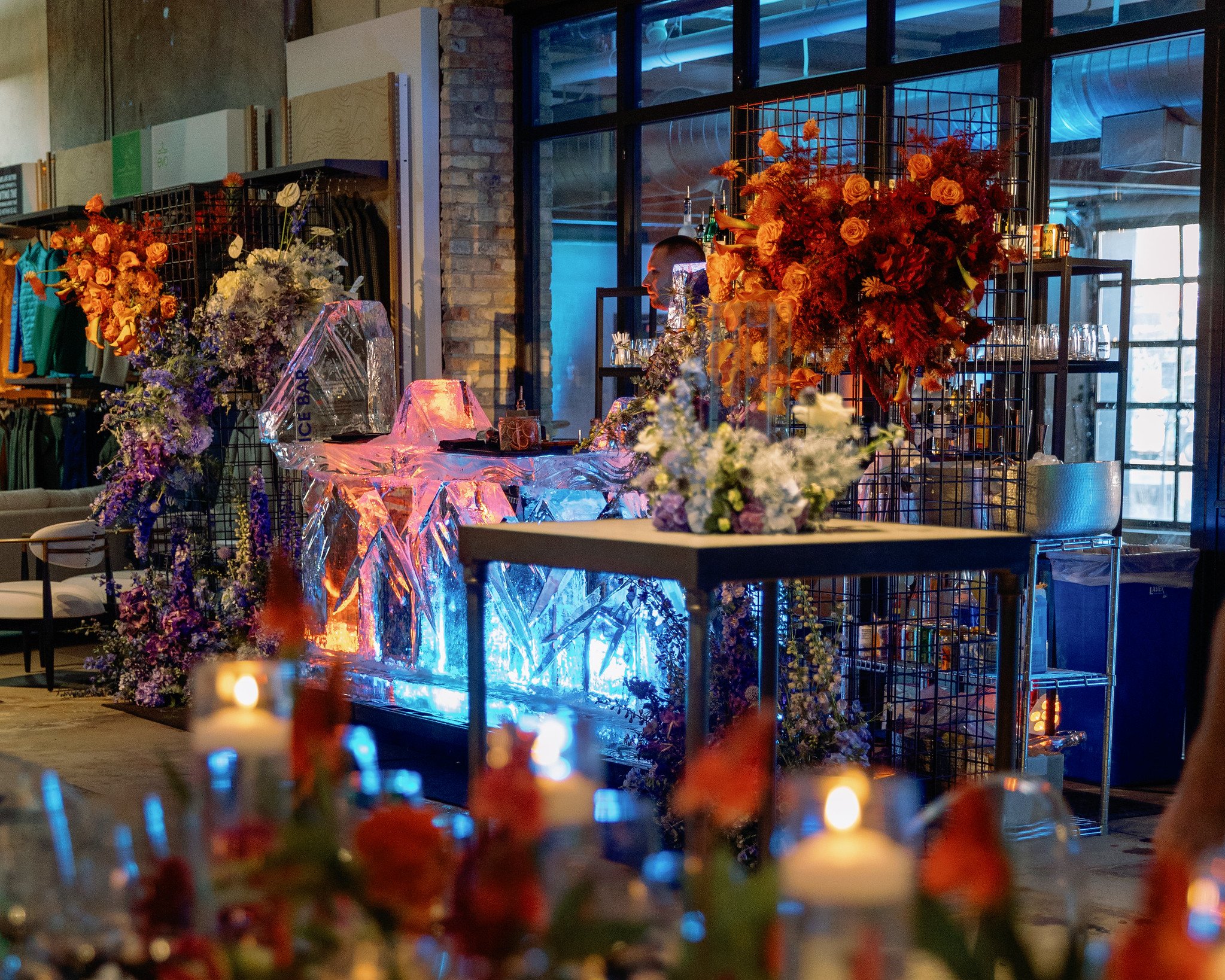 Interior of a modern space decorated with colorful floral arrangements, featuring orange, purple, and white flowers, set near a bar with glasses and bottles, against a brick wall and large windows with a dark urban cityscape outside.