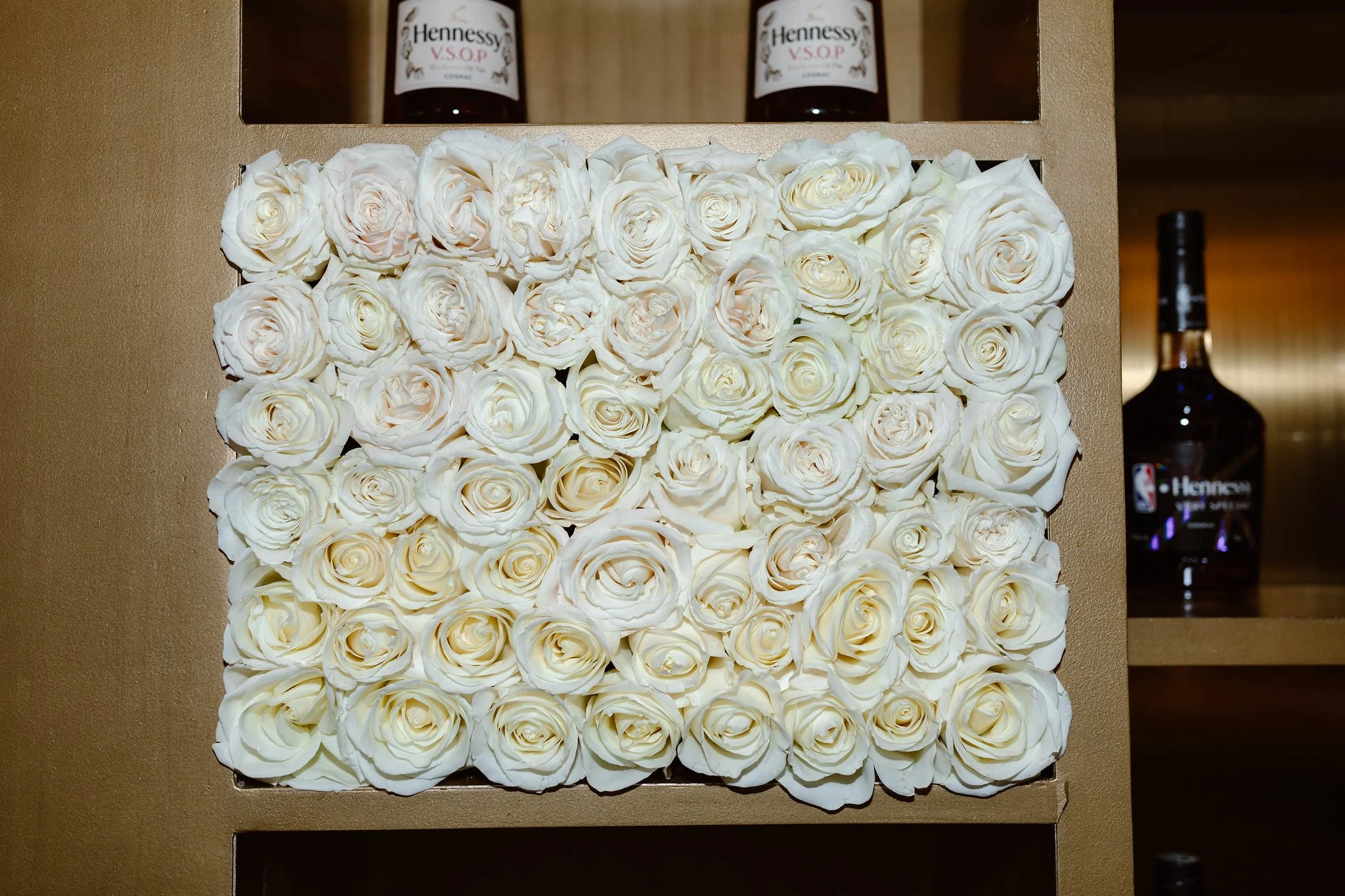 A decorative rectangular arrangement of cream-colored roses. Two bottles of Hennessy V.S.O.P. cognac are on a shelf above the flowers.