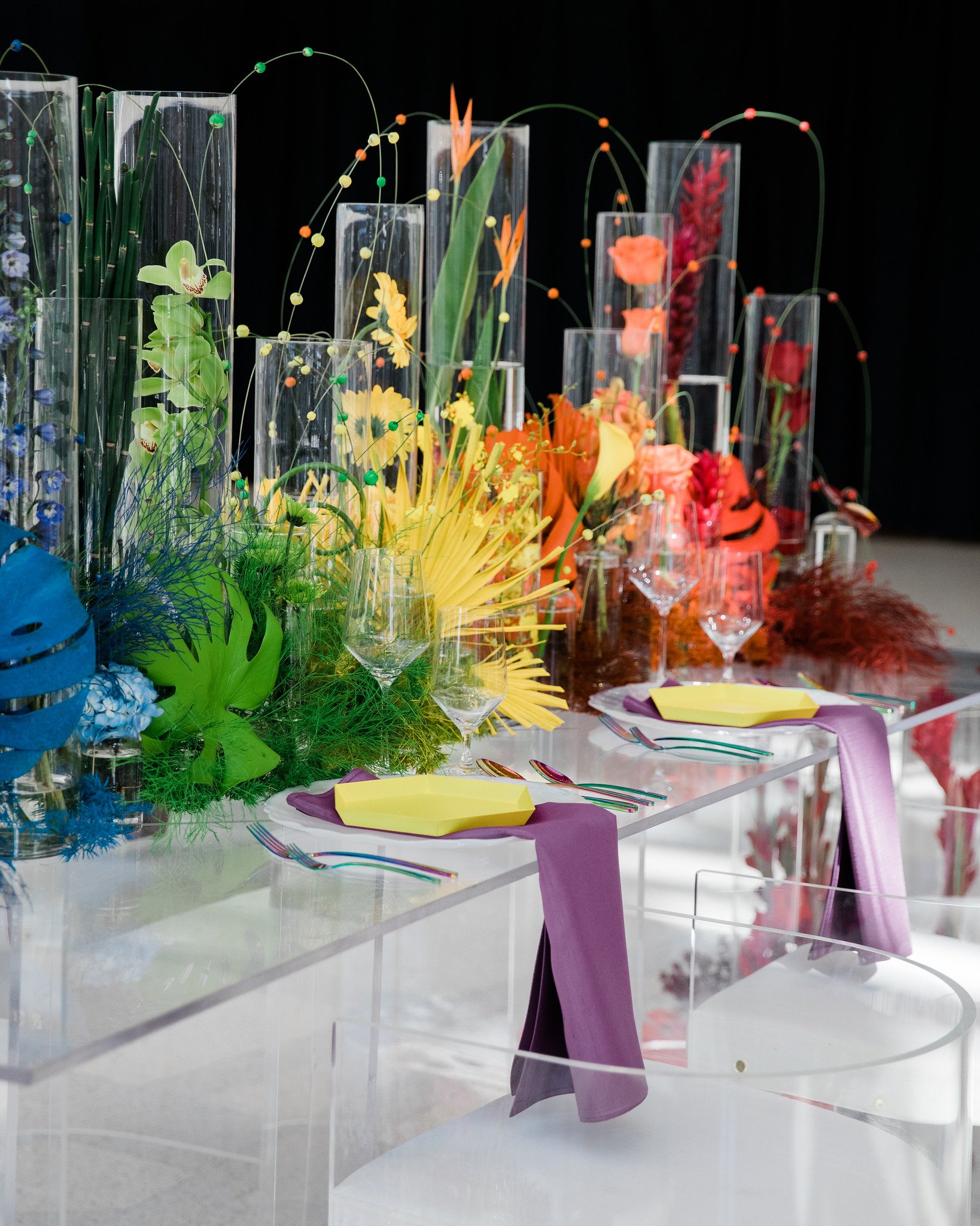 Colorful floral arrangement on a table with purple, yellow, orange, and red flowers in tall glass vases against a black background. Purple and pink cloth napkins with silverware and yellow plates are set on the table.