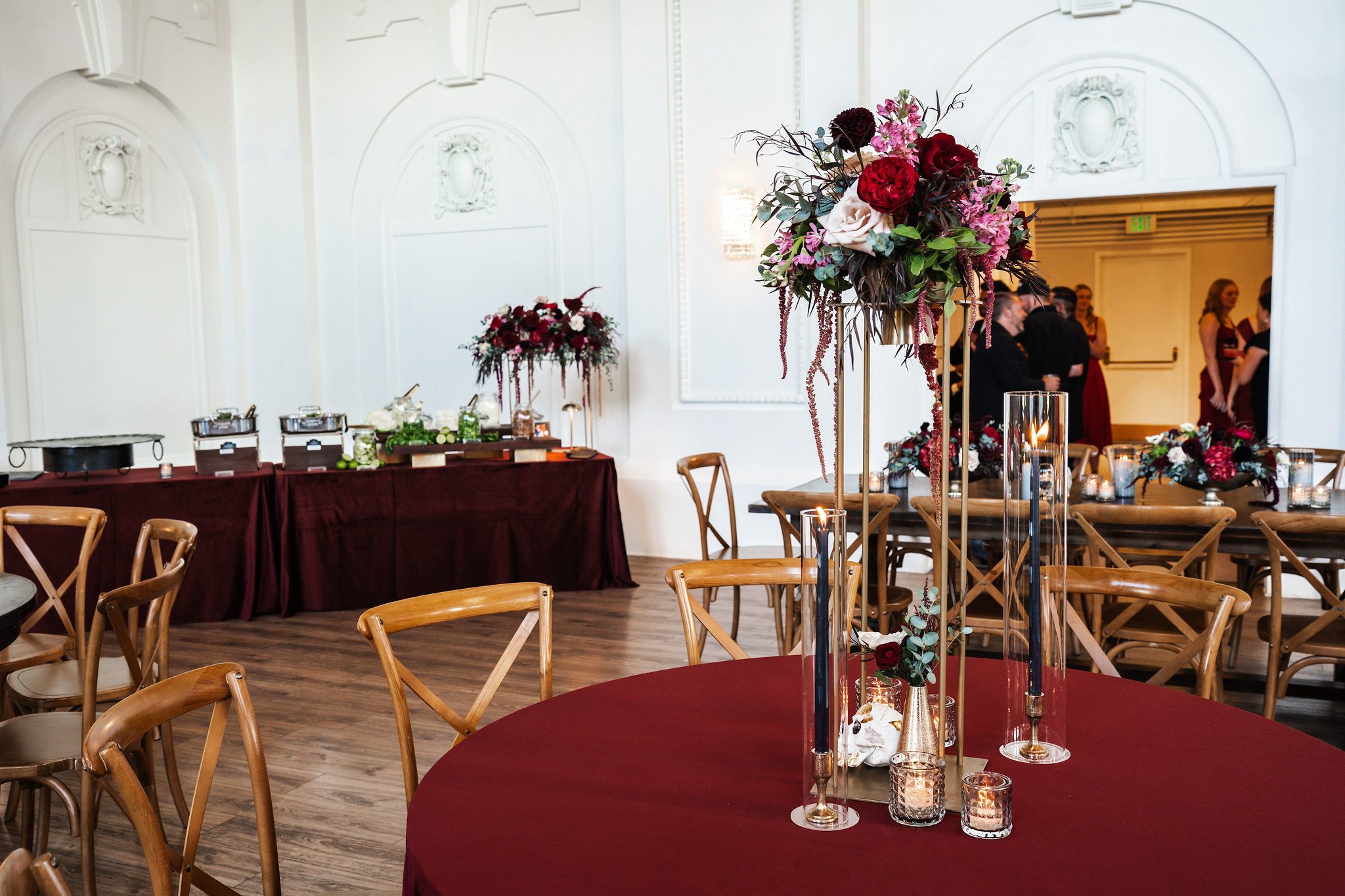 A decorated event space featuring tables with burgundy tablecloths, tall floral centerpieces with red, white, and pink flowers, and candlelit glass holders. In the background, people are gathered near a buffet area with additional floral arrangements