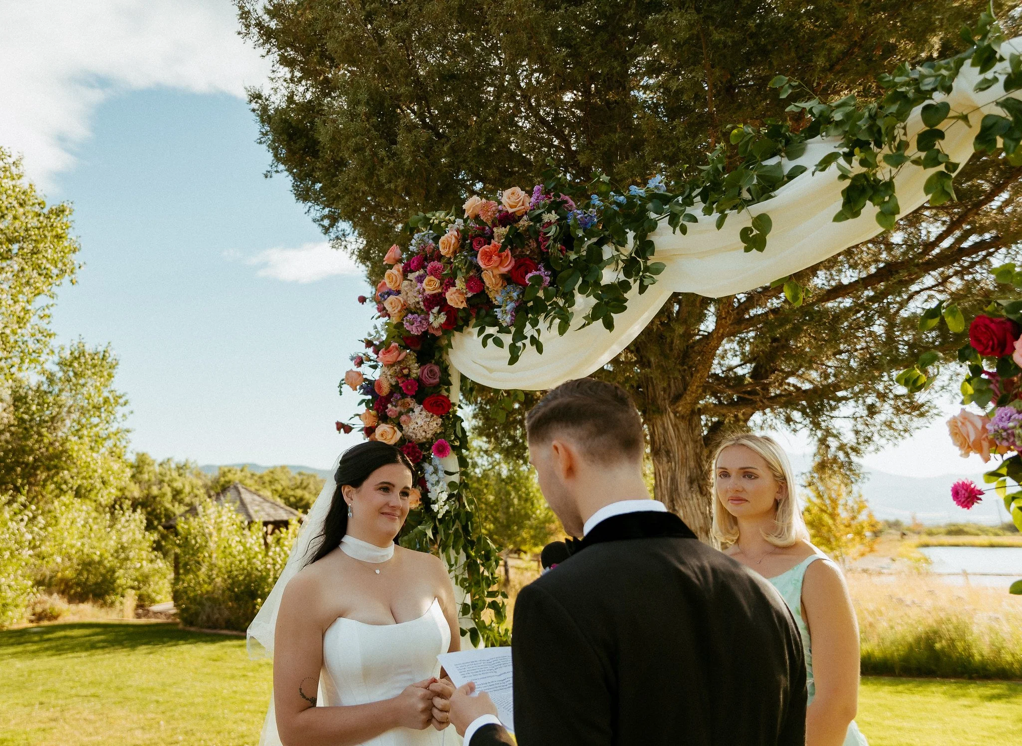 A bride and groom exchanging vows during a wedding ceremony outdoors under a floral arch with trees and blue sky in the background.