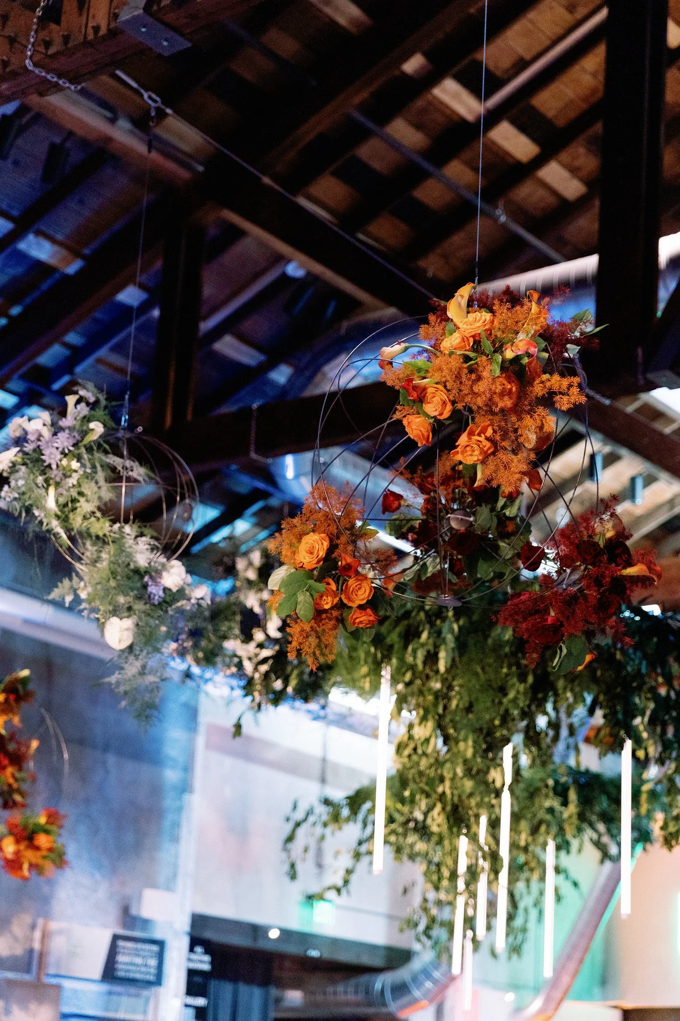 Hanging floral arrangements with orange, purple, and white flowers and green leaves, suspended from a ceiling with dark wooden beams.