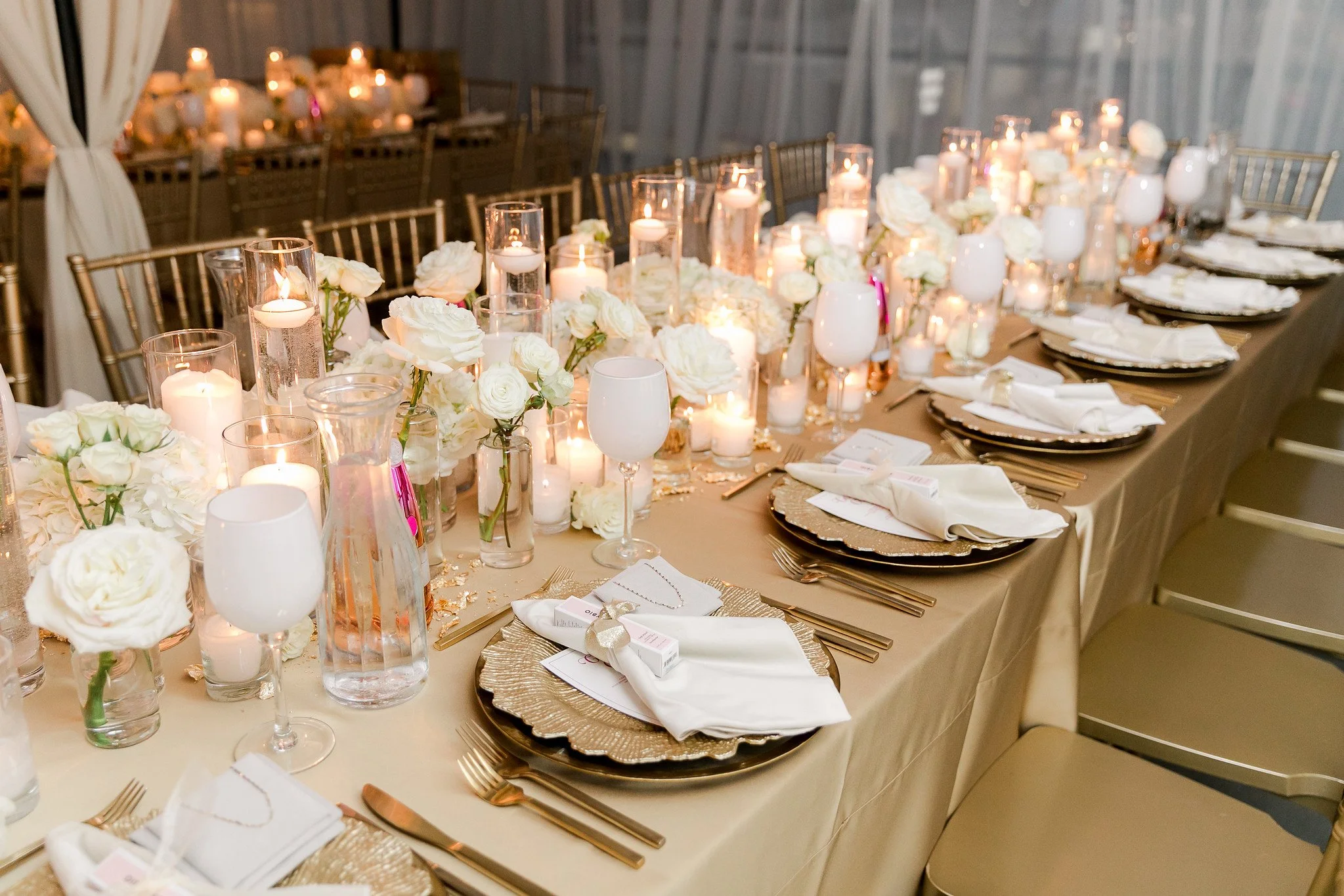 Illuminated table setting at a formal event with white roses and candles arranged along the center, gold placemats, white napkins, and gold utensils for each guest, with gold chairs and a gray wall in the background.