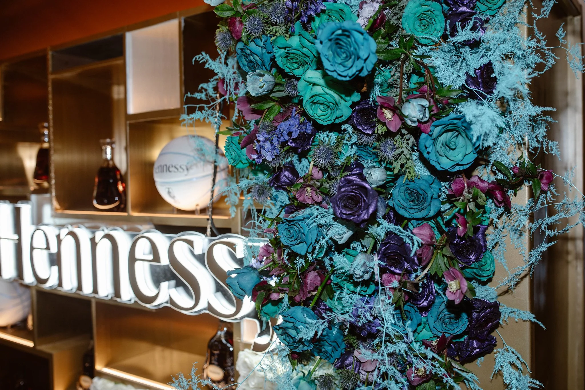 A colorful floral arrangement with blue, purple, and pink flowers and greenery, set against a background of wooden shelving and a neon sign with part of the word "Cheers" visible.