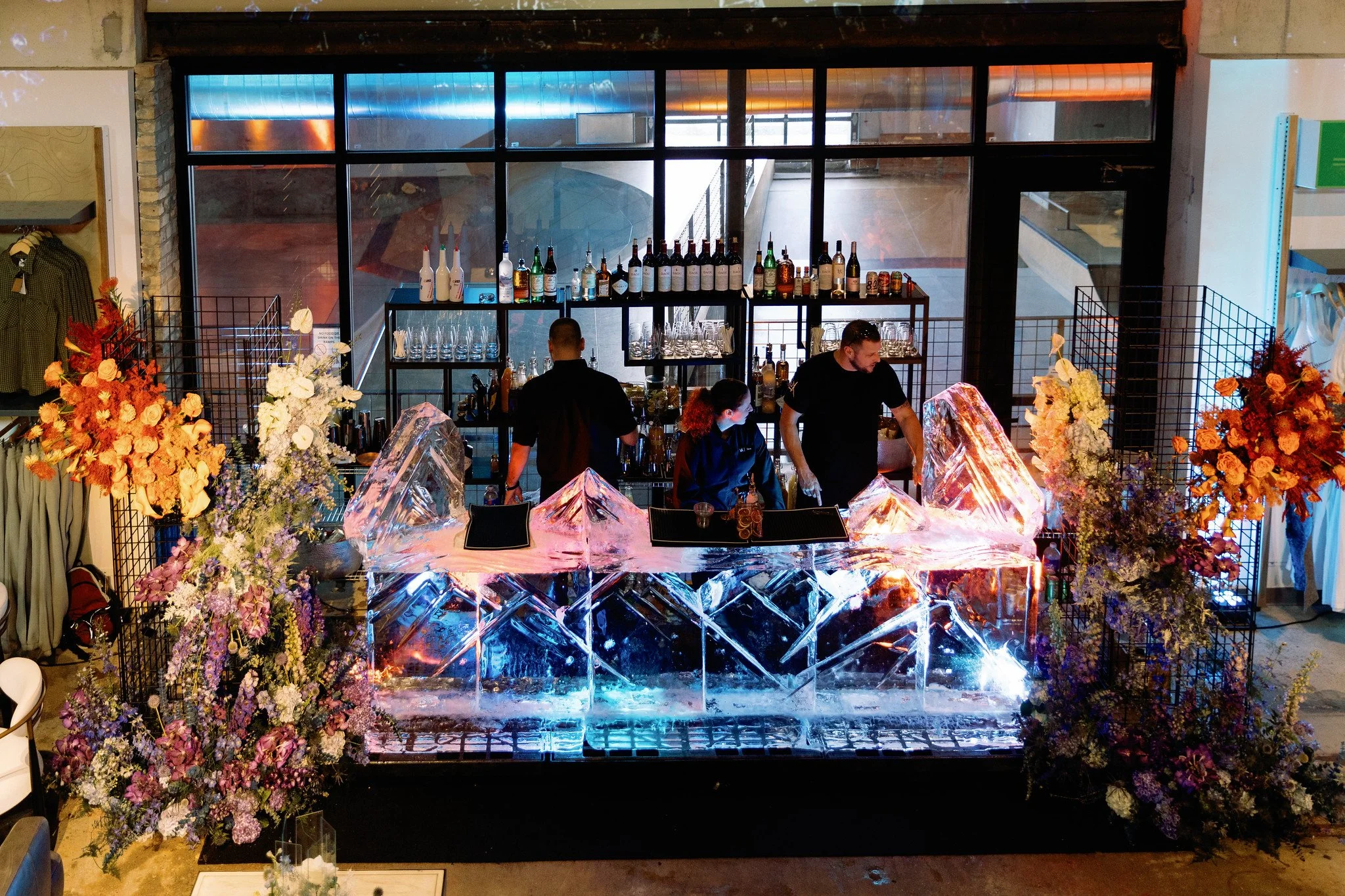 Bar setup with glassware and alcoholic bottles behind ice sculpture, surrounded by floral arrangements