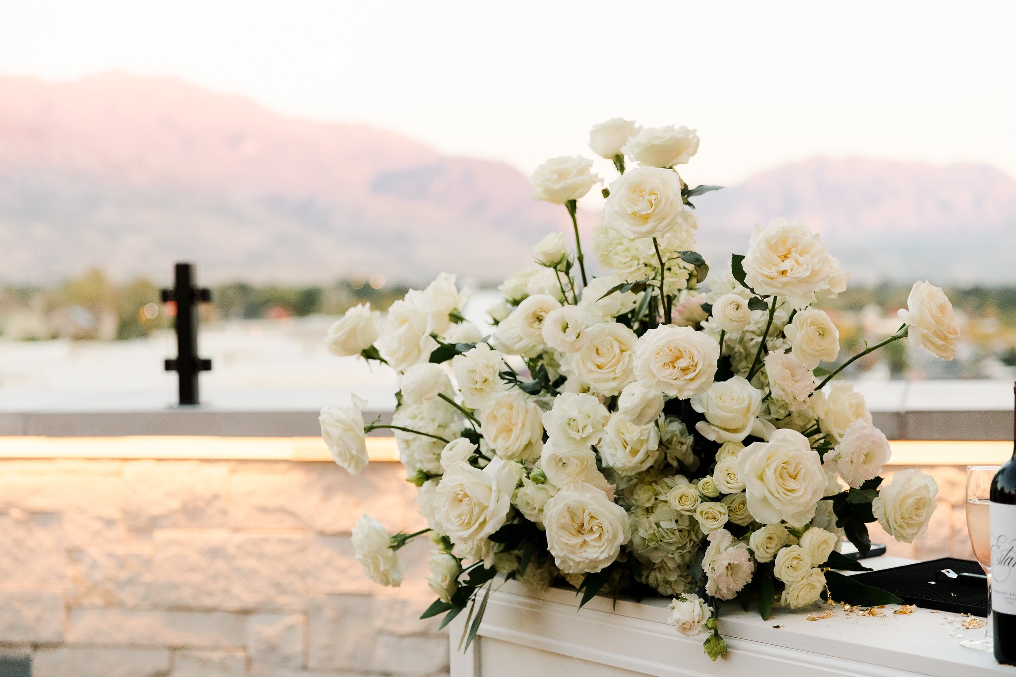 White floral arrangement with roses and other flowers on a table, with a scenic background of distant mountains and a cross in the distance.