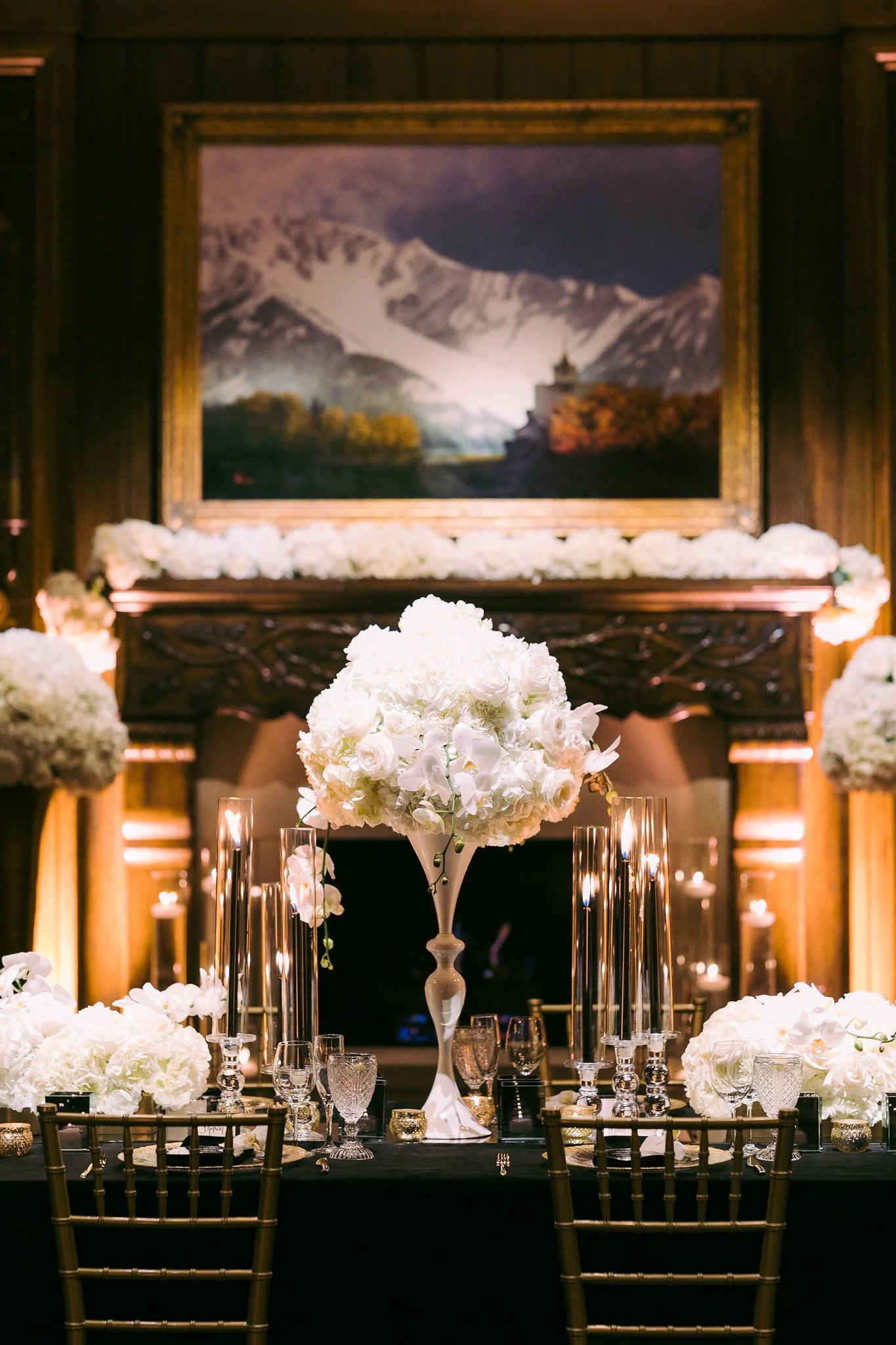 Elegant banquet table with a tall floral centerpiece of white roses and orchids, surrounded by candles and glassware, with a mountain landscape painting hanging on a dark wood paneled wall in the background.