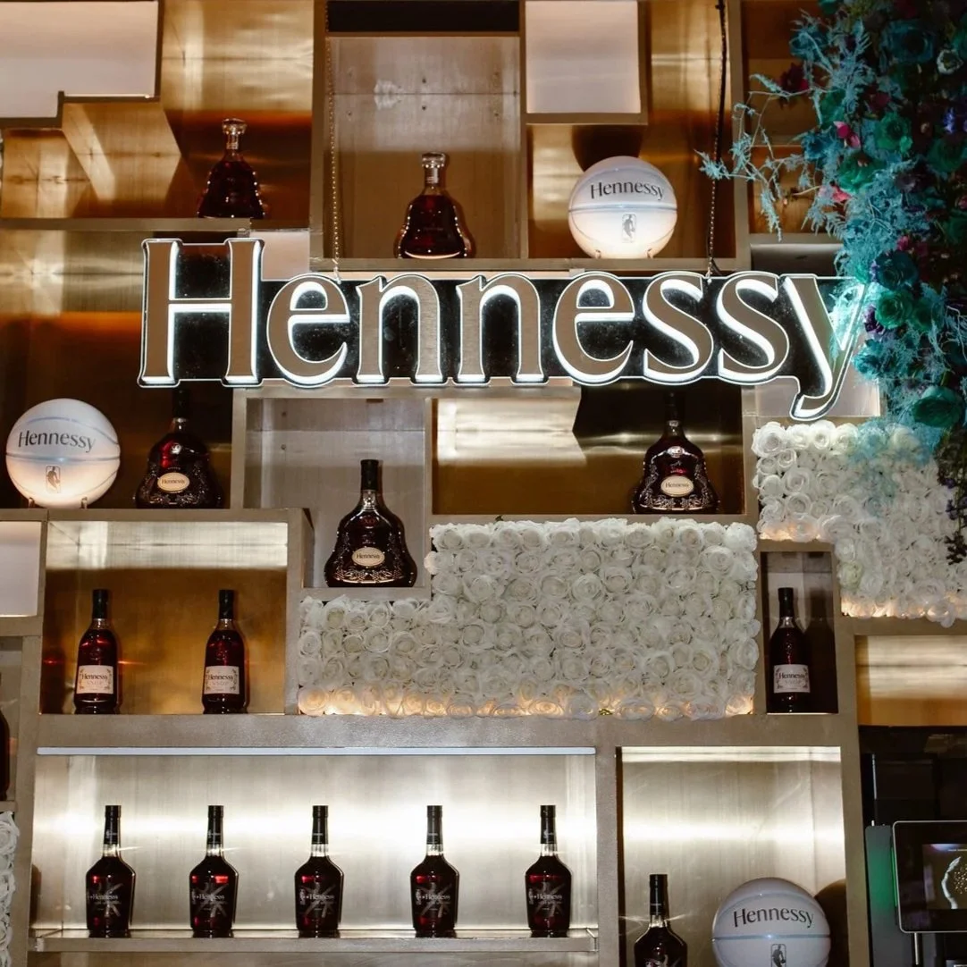 Display of Hennessy bottles and branded decorations behind a illuminated Hennessy sign at an upscale bar or store.