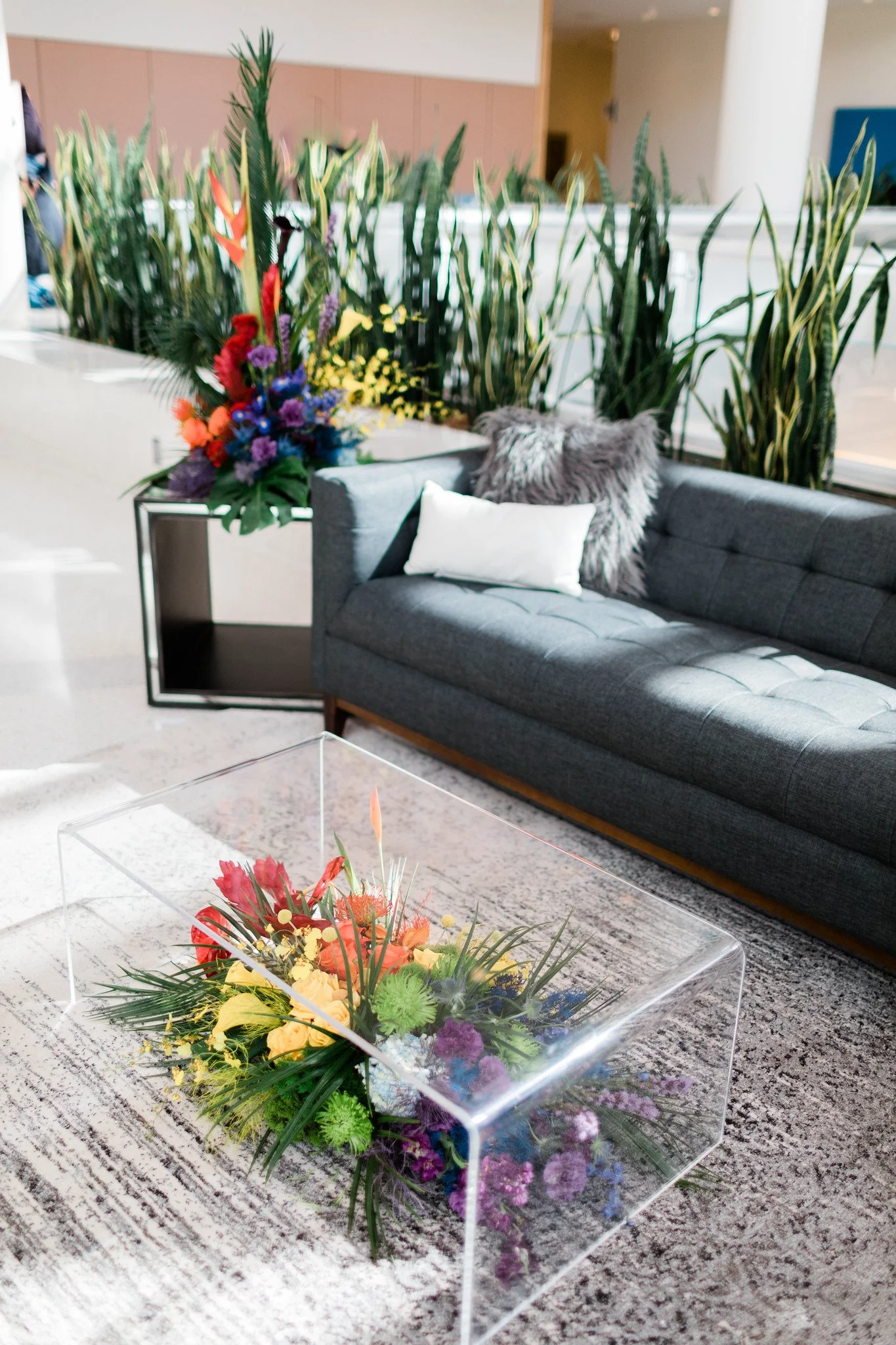 Colorful flower arrangement in a transparent box placed on a patterned area rug in a modern indoor space with gray furniture and tall green plants in the background.